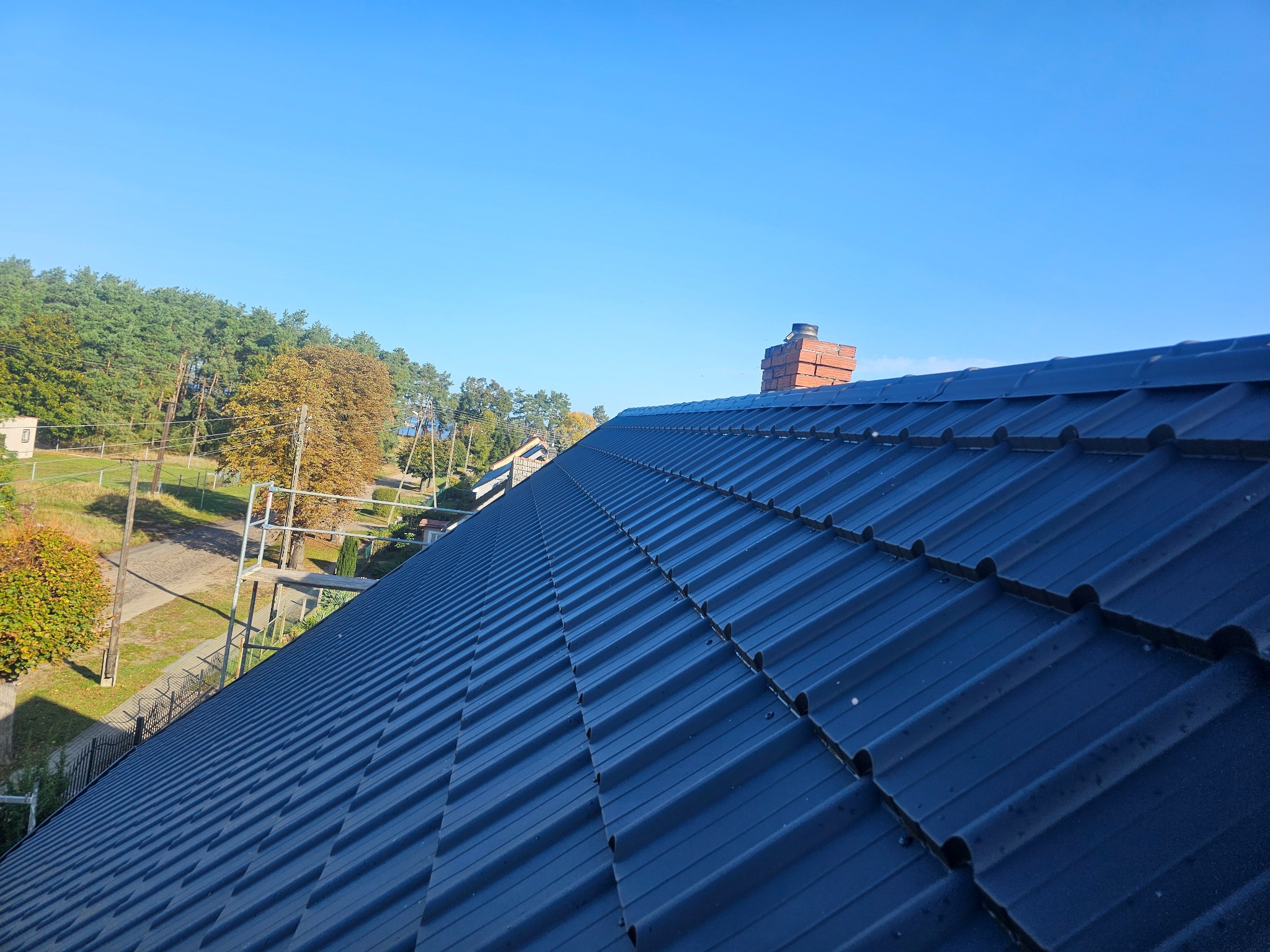 Nowe, ciemne pokrycie dachowe z blachy trapezowej, widok z góry na tle błękitnego nieba i zieleni drzew, z kominem z czerwonej cegły w tle.
