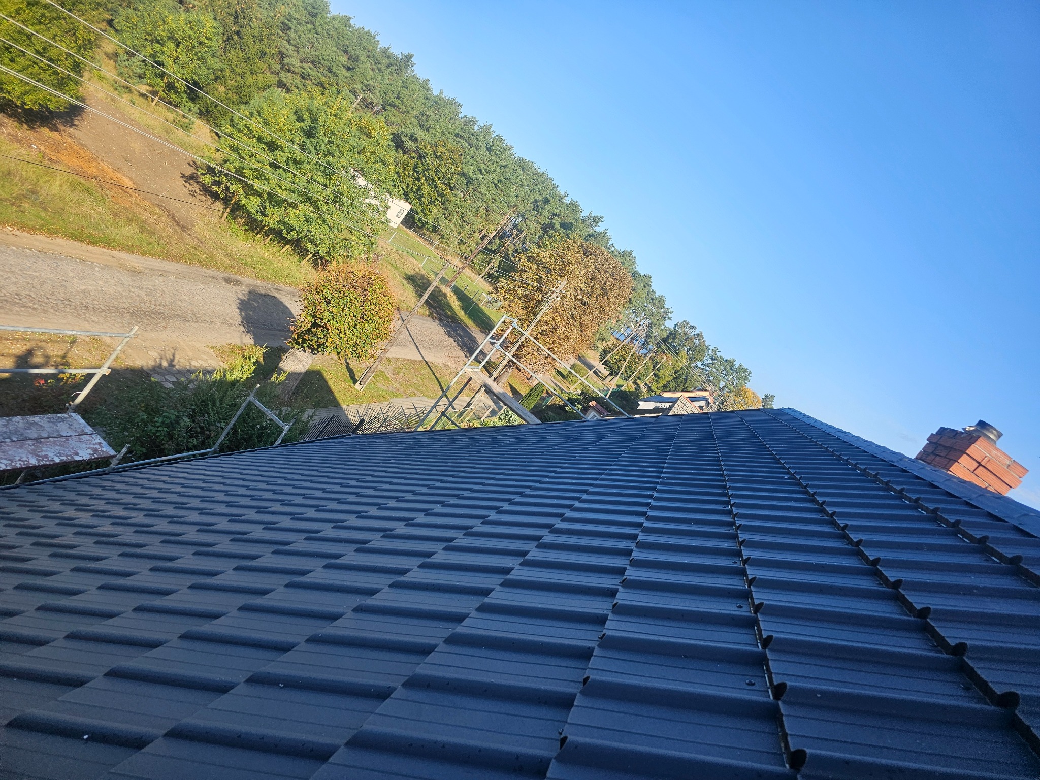 Nowy, ciemny dach z blachodachówki modułowej, widok z góry na tle drzew i błękitnego nieba, rusztowanie w lewym górnym rogu.