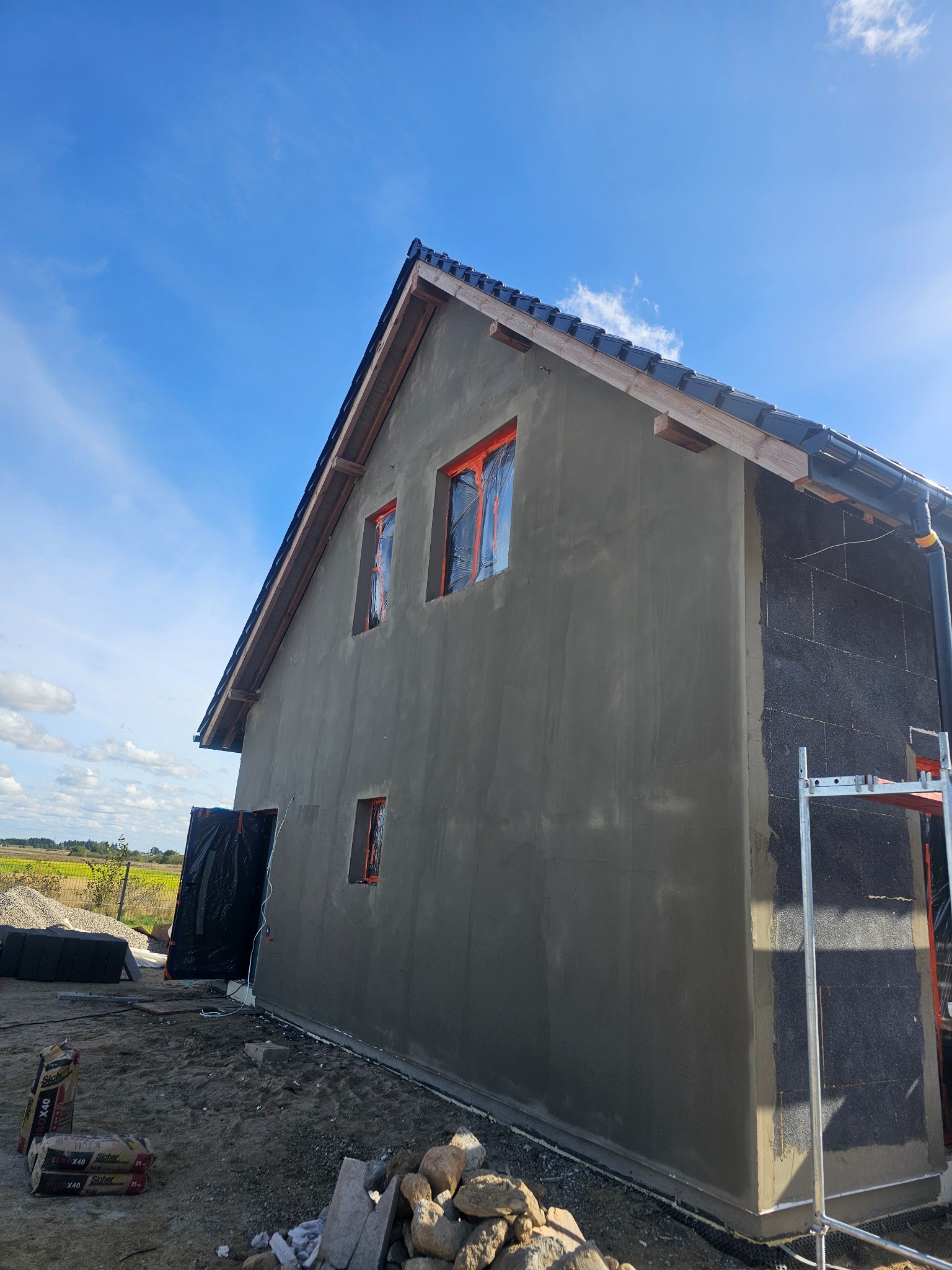 Dom w trakcie prac elewacyjnych: szara, surowa ściana z trzema oknami zabezpieczonymi folią, fragment czarnej izolacji termicznej, rusztowanie oraz worki z zaprawą na ziemi. W tle błękitne niebo.