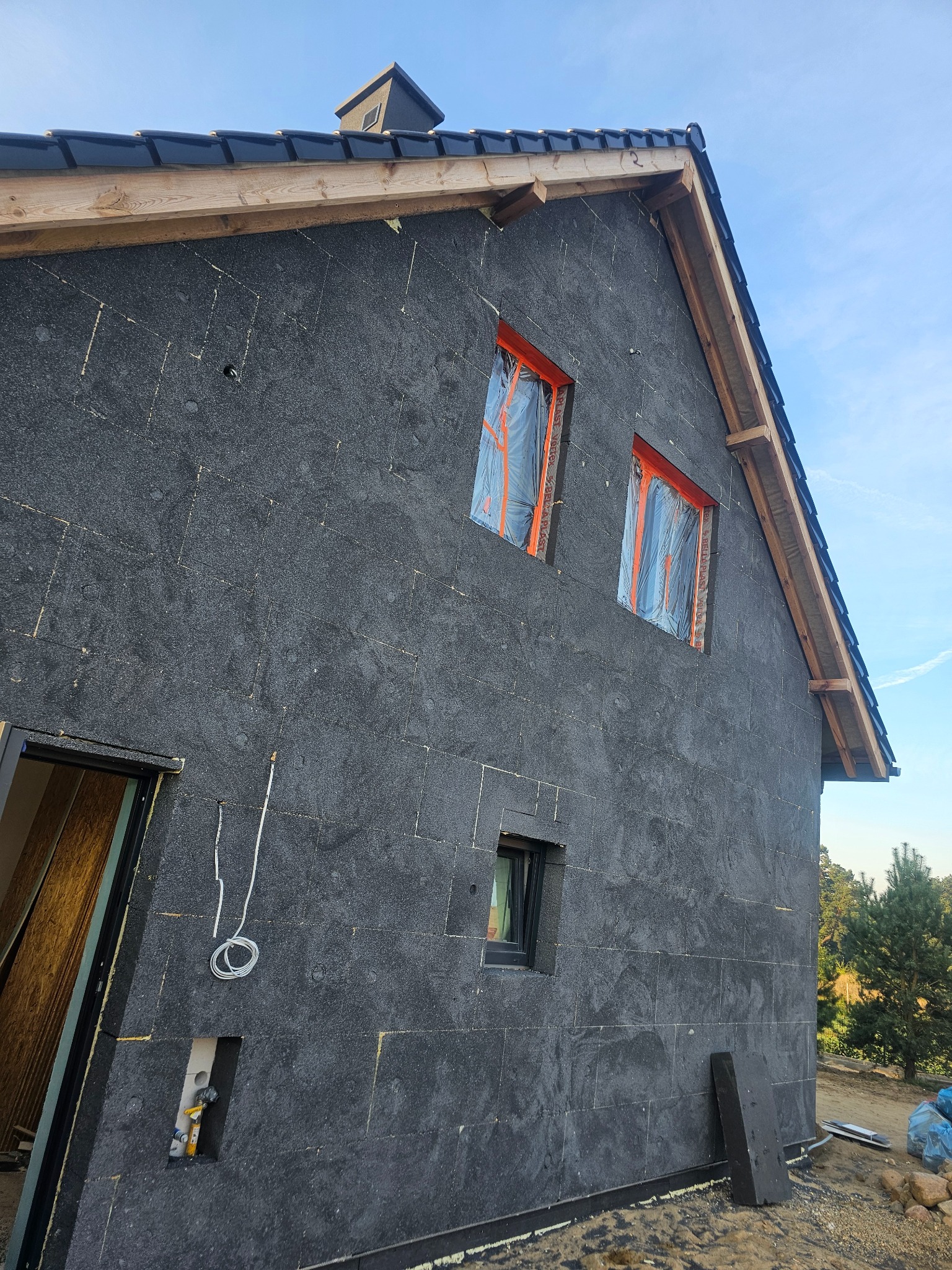 Dom w trakcie ocieplania elewacji, widoczne czarne płyty izolacyjne, zabezpieczone okna folią i fragment nowo położonego dachu z czarnej dachówki ceramicznej.