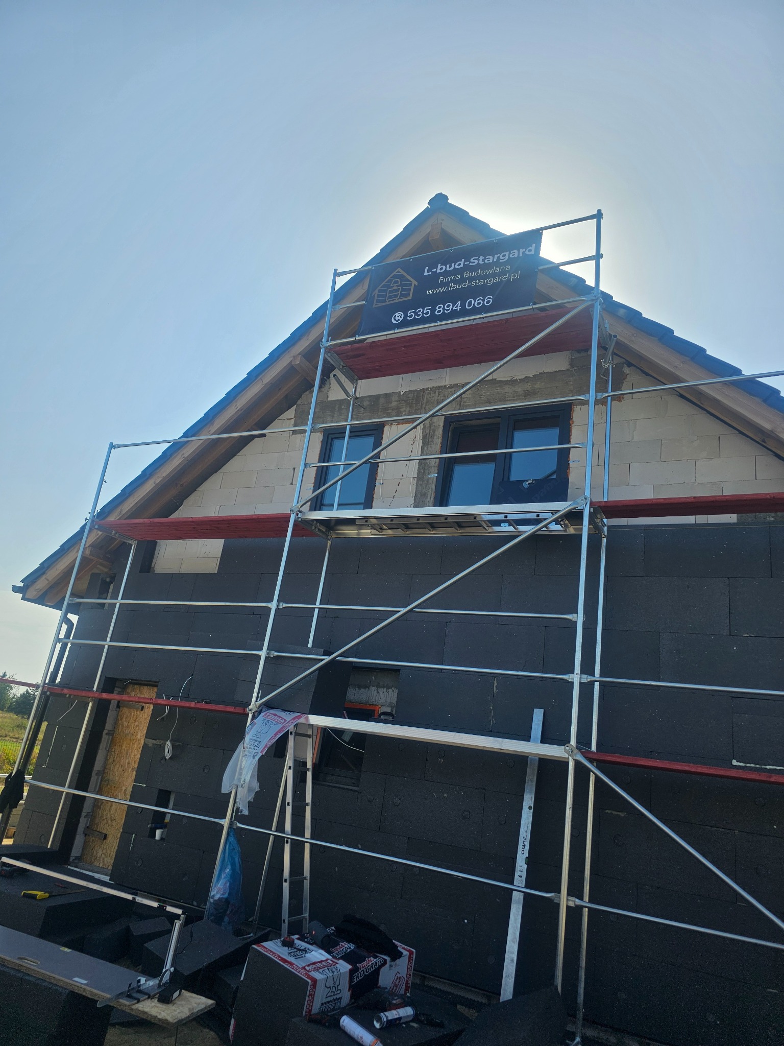 Dom w trakcie ocieplania elewacji, widoczne rusztowanie, czarne płyty izolacyjne, okna i fragment dachu z szarą dachówką. Na rusztowaniu baner reklamowy firmy budowlanej L-bud-Stargard.