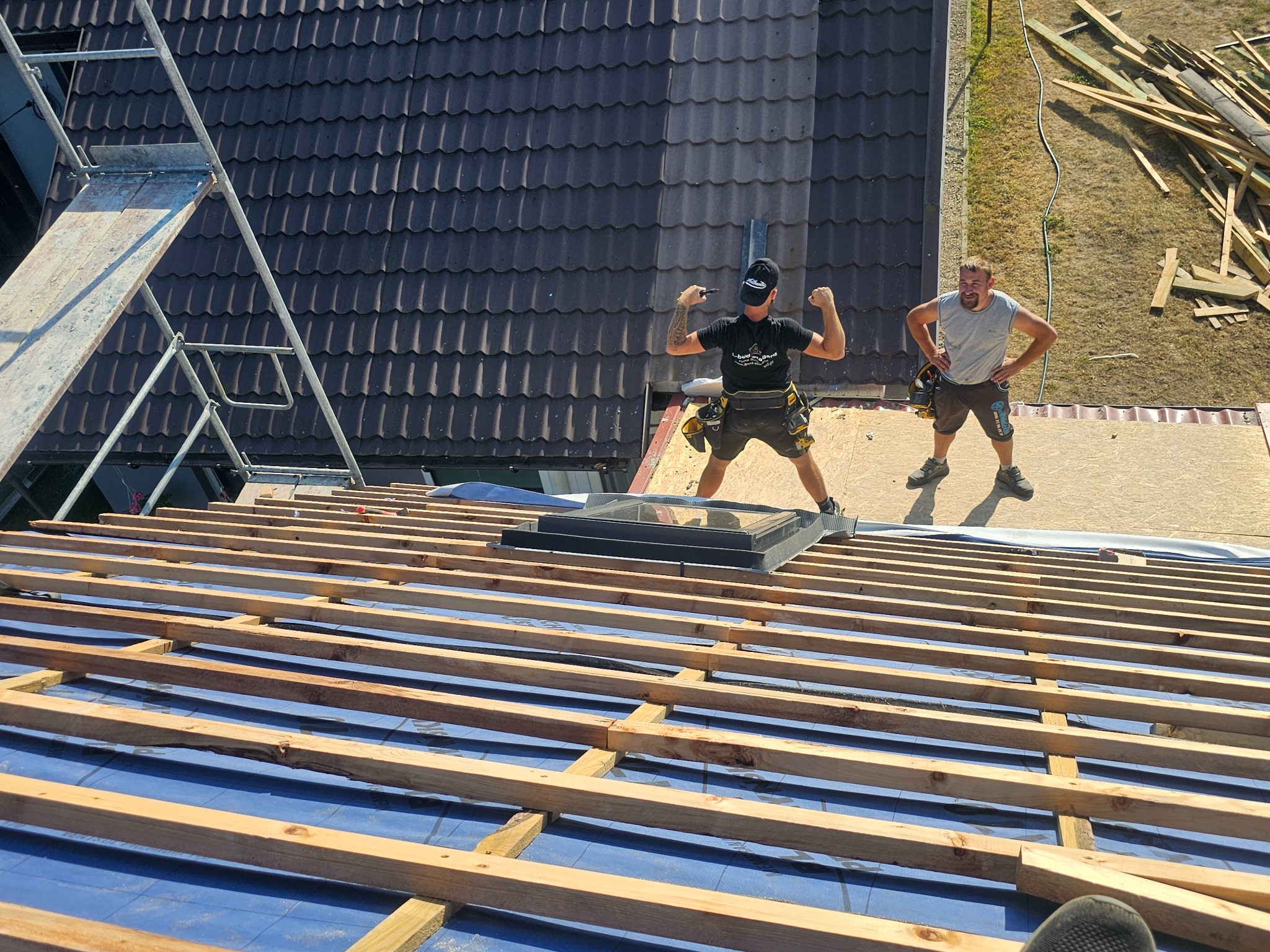 Ekipa dekarzy na dachu z zamontowanym oknem dachowym, jeden z nich w geście triumfu prezentuje bicepsy, w tle widoczna sterta drewna i gotowy fragment dachu z dachówką.