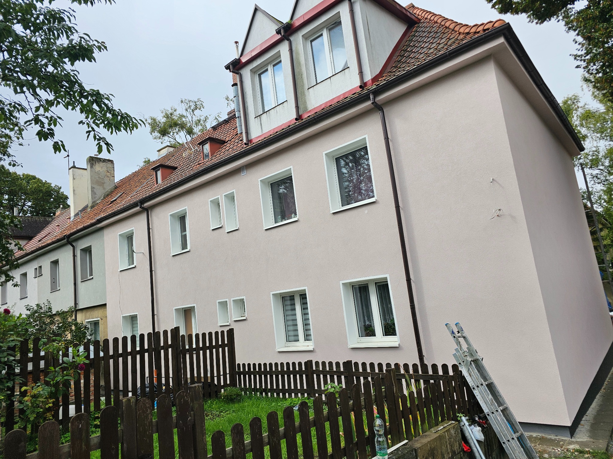 Odnowiona elewacja dwupiętrowego budynku z nową, jasnoróżową fasadą, widoczna drabina oparta o ścianę i drewniane ogrodzenie.