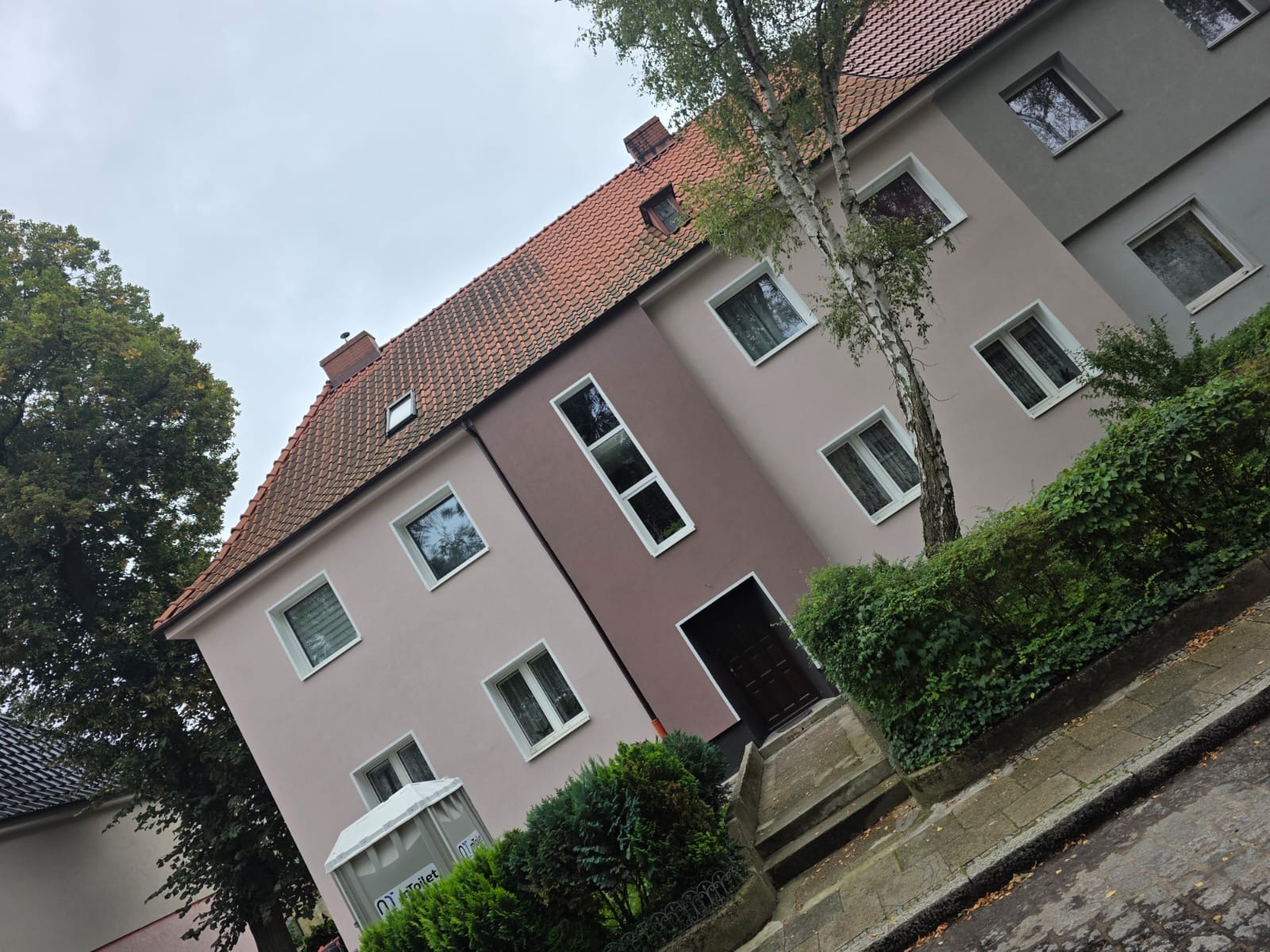 Odnowiona elewacja dwukondygnacyjnego budynku z czerwonym dachem, z widocznymi oknami i zielenią. Uwagę zwraca różnica kolorów między segmentami elewacji.