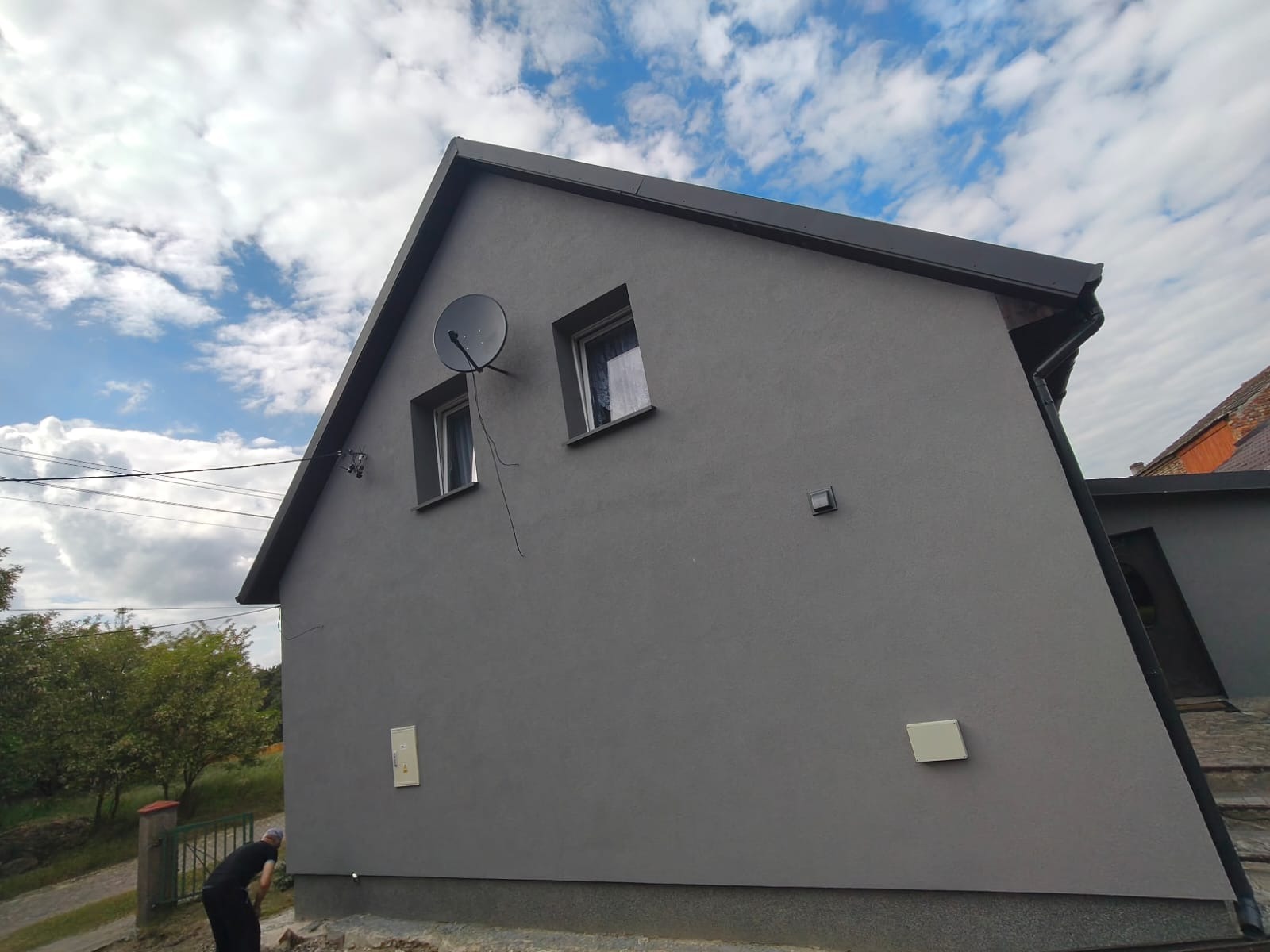 Dom z szarą elewacją w słoneczny dzień. Widoczne dwa okna, antena satelitarna i fragment dachu. Na dole kadru osoba wykonująca prace ziemne.