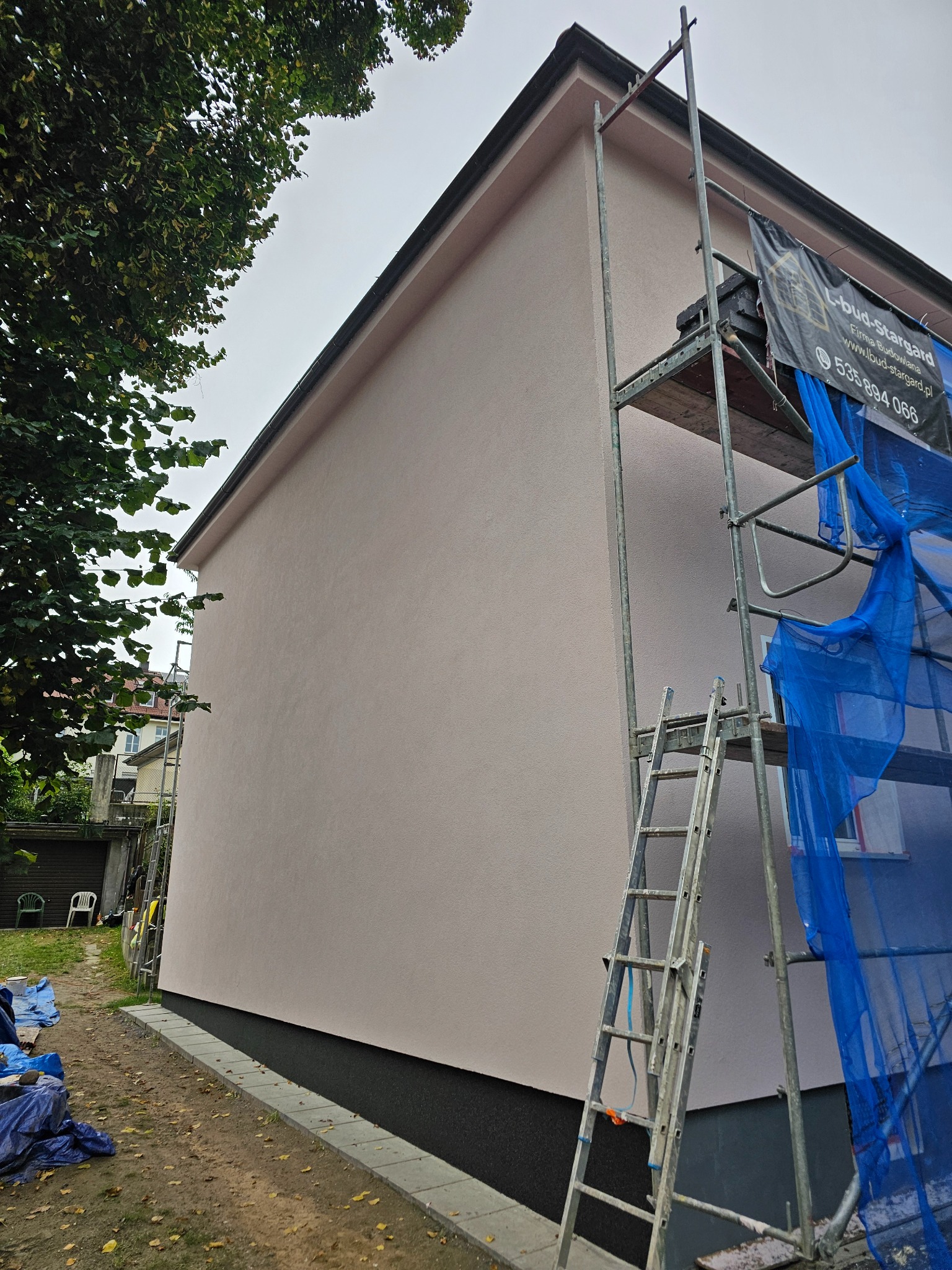 Świeżo pomalowana elewacja budynku w jasnym odcieniu różu, z widocznym fragmentem rusztowania i banerem reklamowym firmy budowlanej, na tle zieleni i pochmurnego nieba.