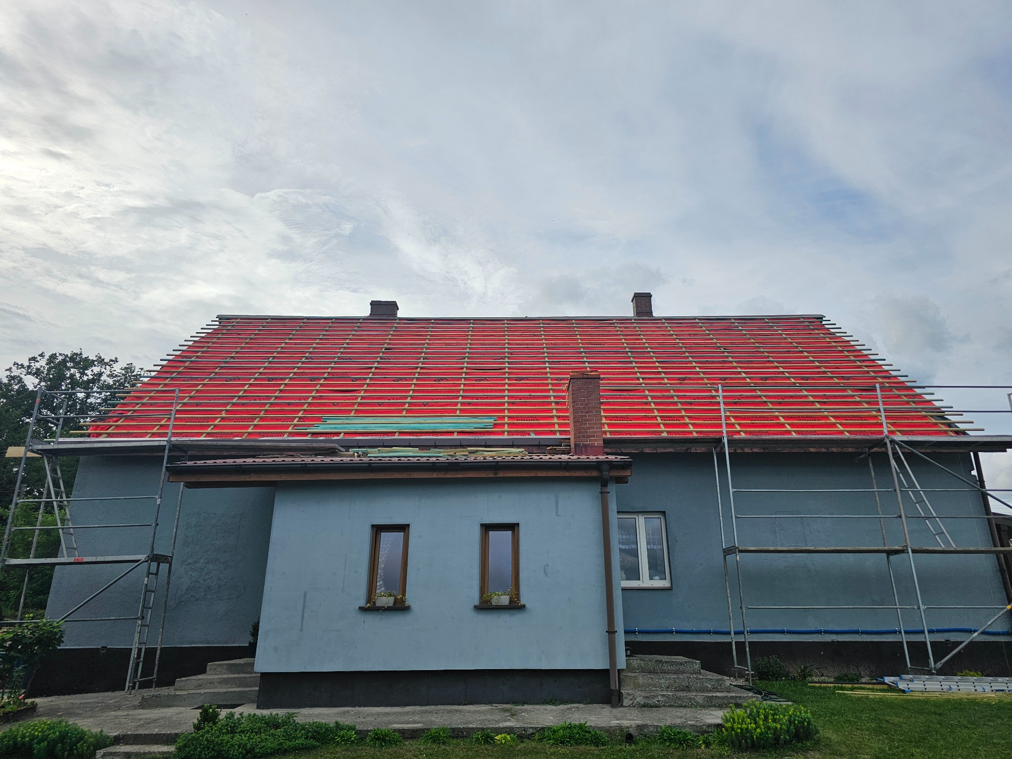 Dom z częściowo zdjętym starym dachem i przygotowanym podkładem z czerwonej membrany dachowej. Widoczne rusztowania i dwa kominy. Etap prac przygotowawczych.