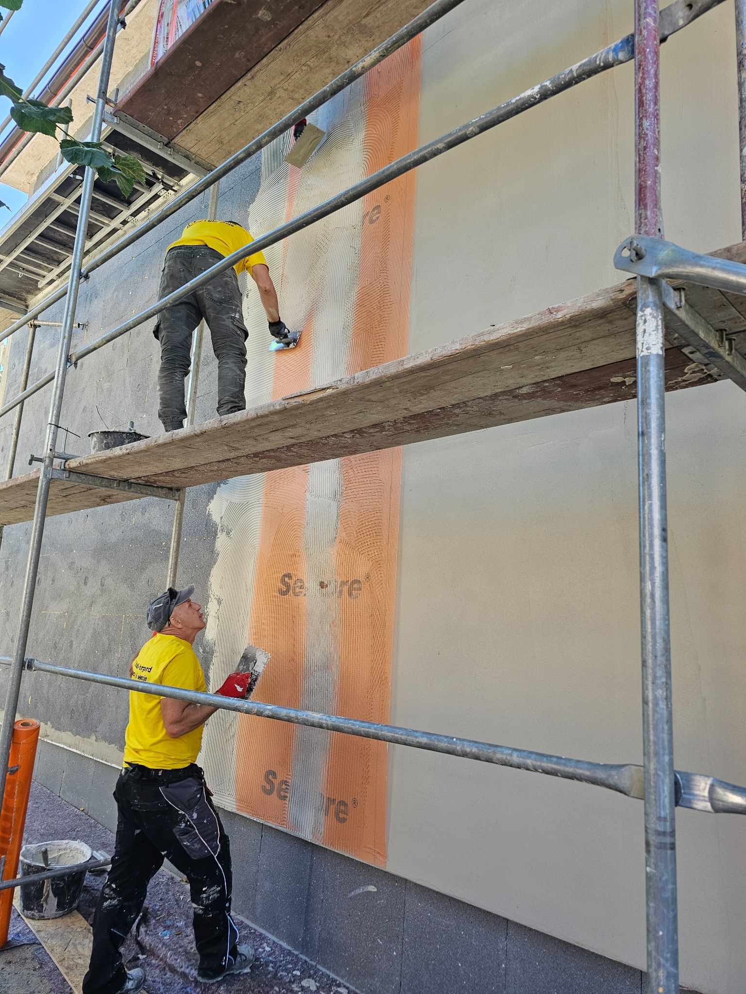 Dwóch pracowników na rusztowaniu aplikuje zaprawę na ocieploną ścianę budynku. Widoczne narzędzia i materiały budowlane. Etap prac wykończeniowych elewacji.