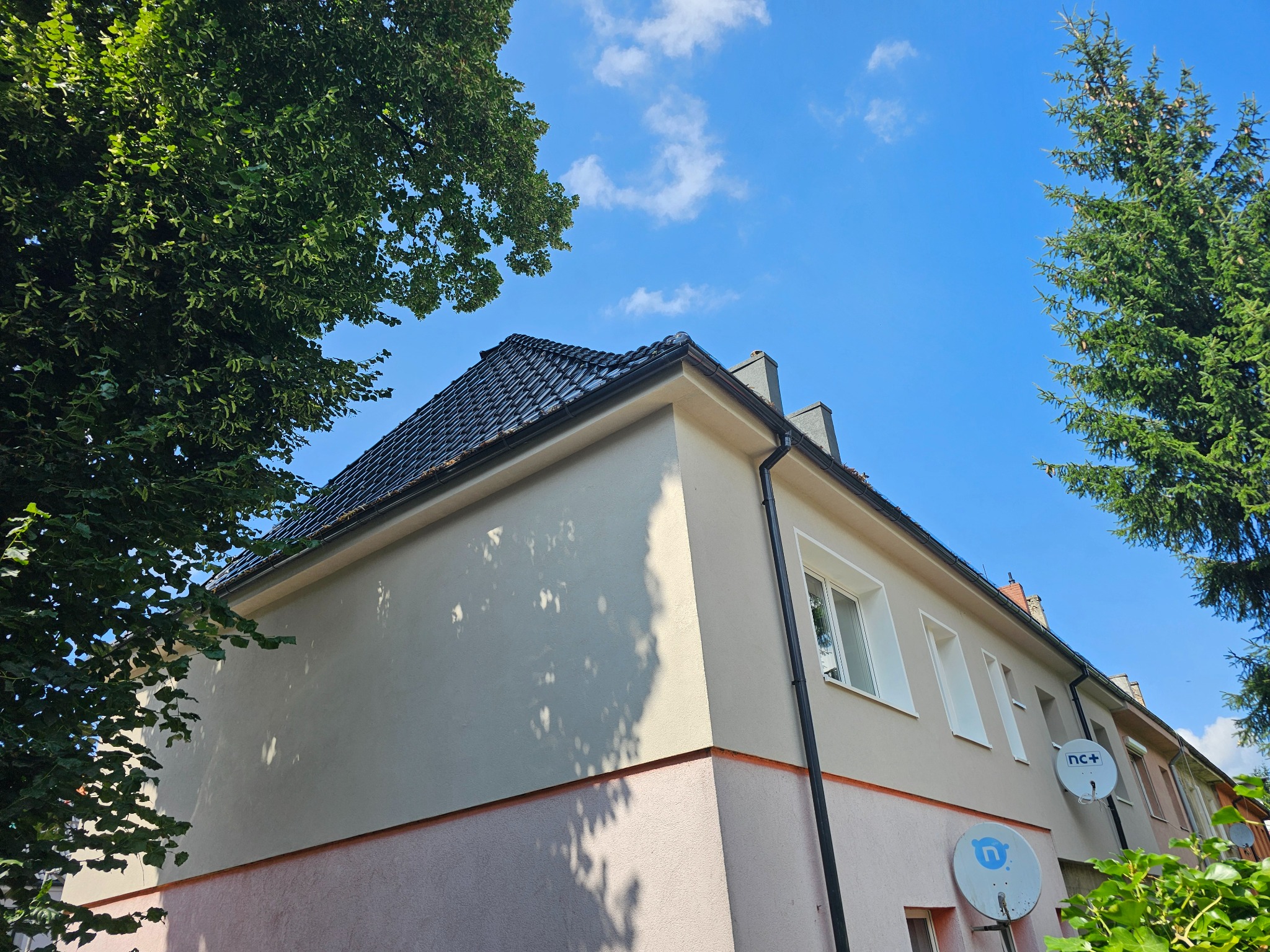 Dwukondygnacyjny dom z nowym, czarnym dachem z dachówki, beżową elewacją i antenami satelitarnymi na tle błękitnego nieba i zieleni drzew.