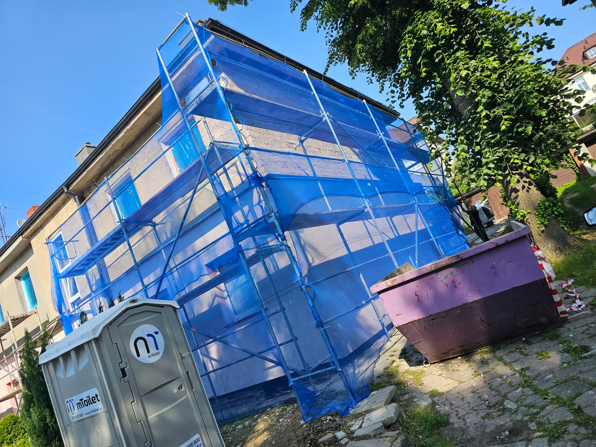 Dom w rusztowaniach z niebieską siatką, widoczny kontener i toaleta przenośna. Przygotowania do prac elewacyjnych lub dekarskich w słoneczny dzień.