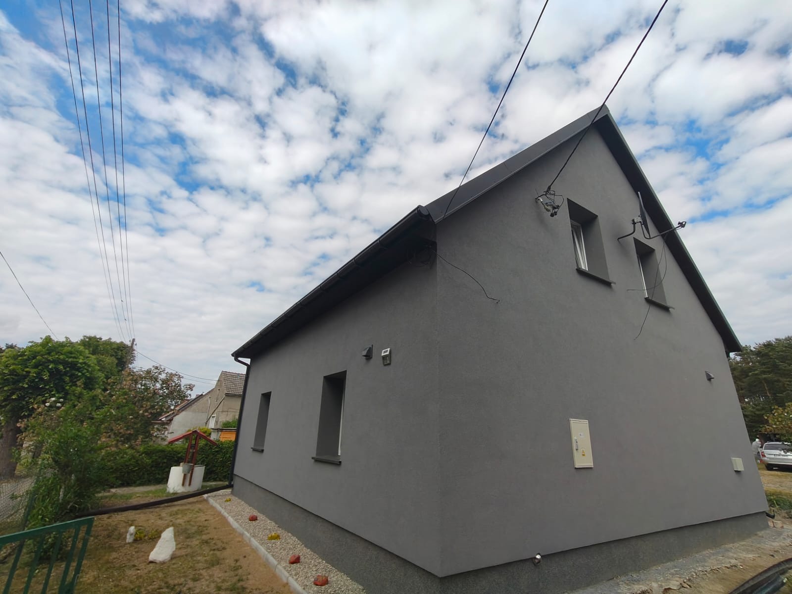 Szary dom jednorodzinny po renowacji elewacji, widok z boku, z czarnym dachem i elementami instalacji na ścianie, na tle błękitnego nieba z chmurami.