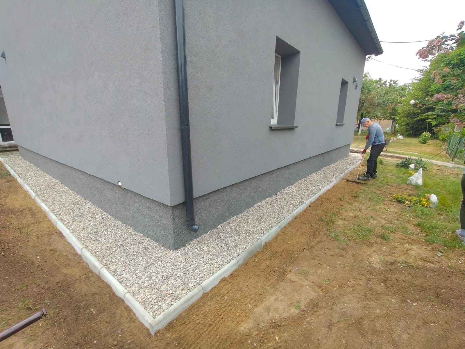 Szara elewacja domu z wykończonym cokołem i kamiennym obramowaniem. Mężczyzna grabi ziemię obok budynku. Widok z niskiego kąta.