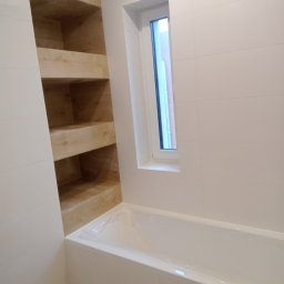 RomBud - Biała łazienka z wanną, oknem i drewnianą wnęką z półkami, wykończona jasnymi płytkami.