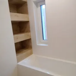 Biała łazienka z wanną, oknem i drewnianą wnęką z półkami, wykończona jasnymi płytkami.