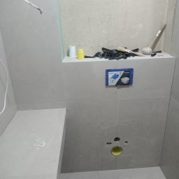 RomBud - Montaż stelaża podtynkowego Geberit Sigma w łazience wykończonej szarymi płytkami, widoczne narzędzia glazurnicze, poziomica, gumowy młotek i elementy dystansowe na półce nad stelażem.