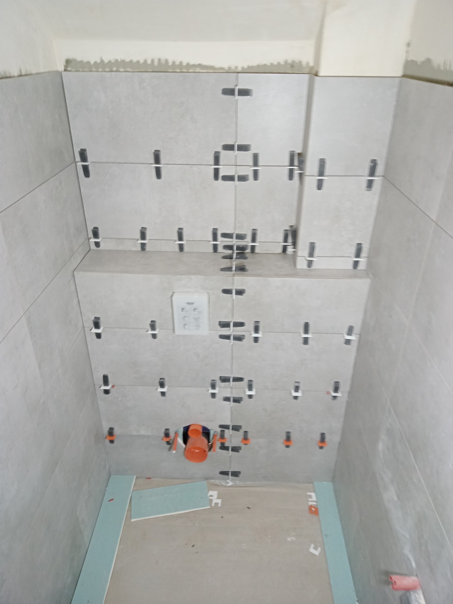 Układanie jasnoszarych płytek w łazience, widoczne krzyżyki dystansowe, rury kanalizacyjne i fragment płyty gipsowo-kartonowej na podłodze.