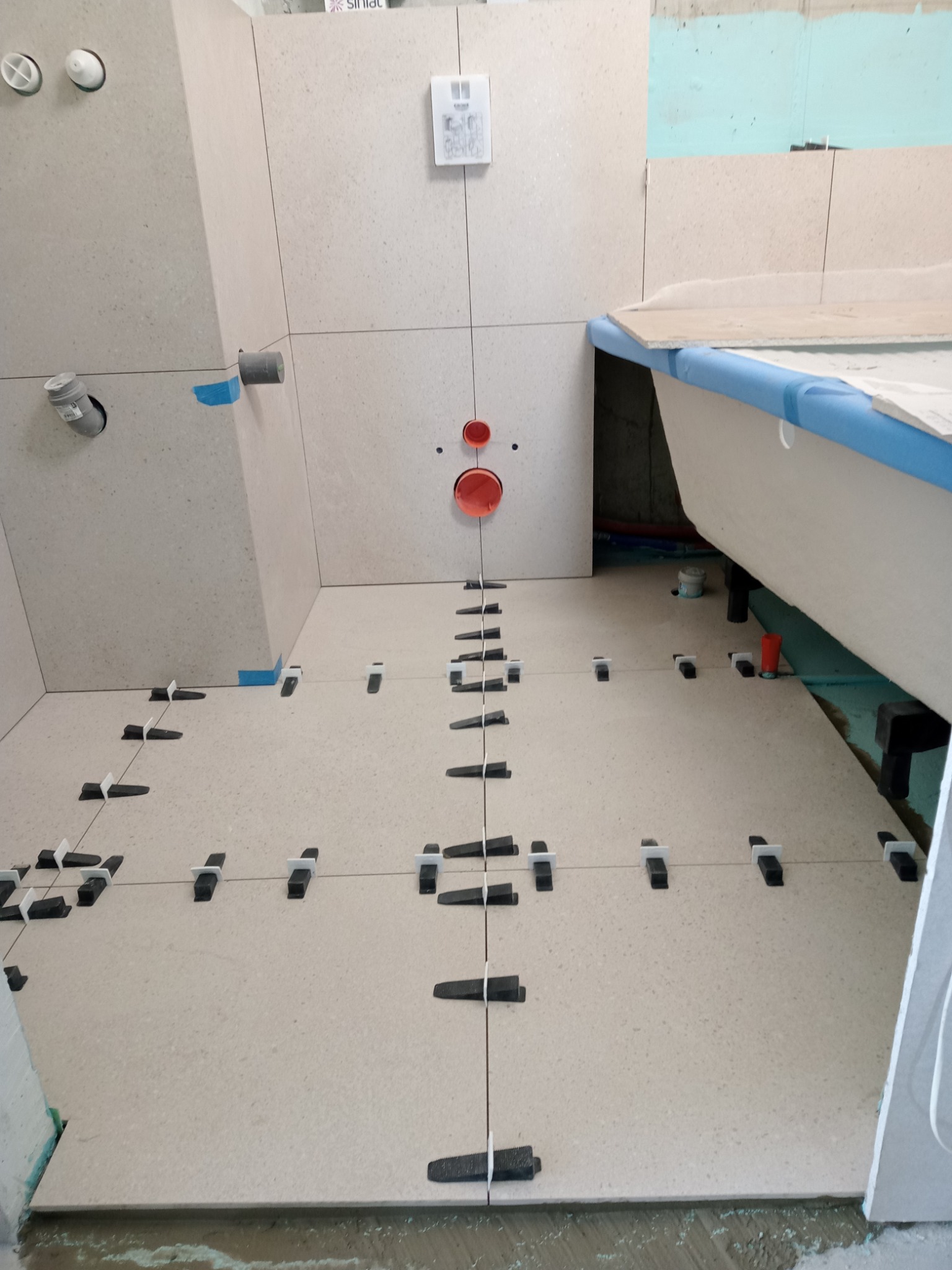 Jasne płytki podłogowe w trakcie układania w łazience, z widocznymi krzyżykami dystansowymi i poziomymi liniami, fragment wanny z niebieskim zabezpieczeniem brzegów w tle, przygotowane otwory na rury.