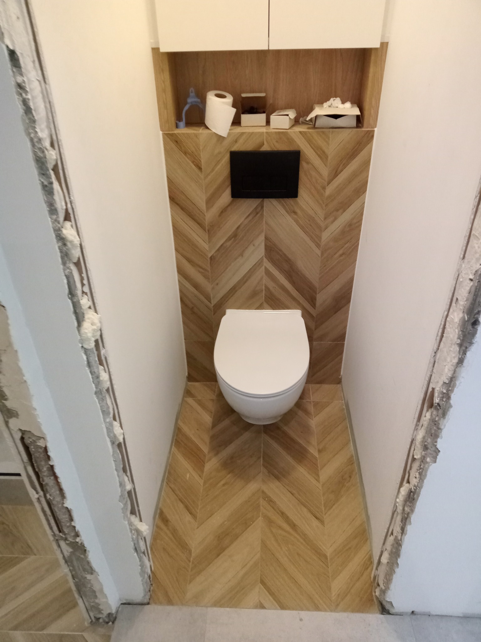 Wisząca toaleta w małej łazience z drewnopodobną glazurą ułożoną w jodełkę na ścianie i podłodze, białe szafki wiszące nad toaletą, ślady pianki montażowej na ościeżnicy po lewej stronie.