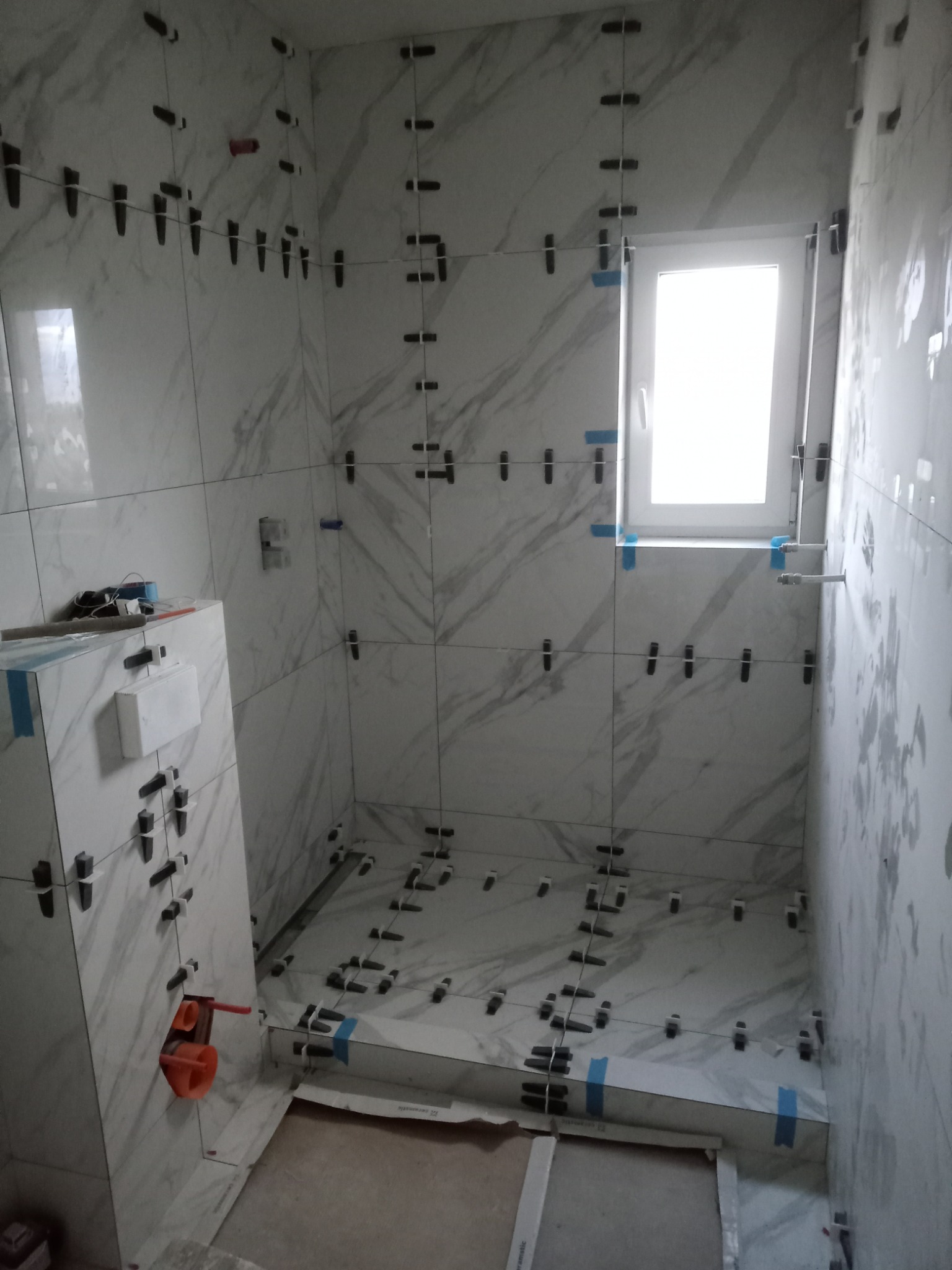 Łazienka w trakcie układania białych płytek imitujących marmur, z użyciem klipsów poziomujących, widoczne fragmenty instalacji hydraulicznej i okno z niebieską taśmą zabezpieczającą.