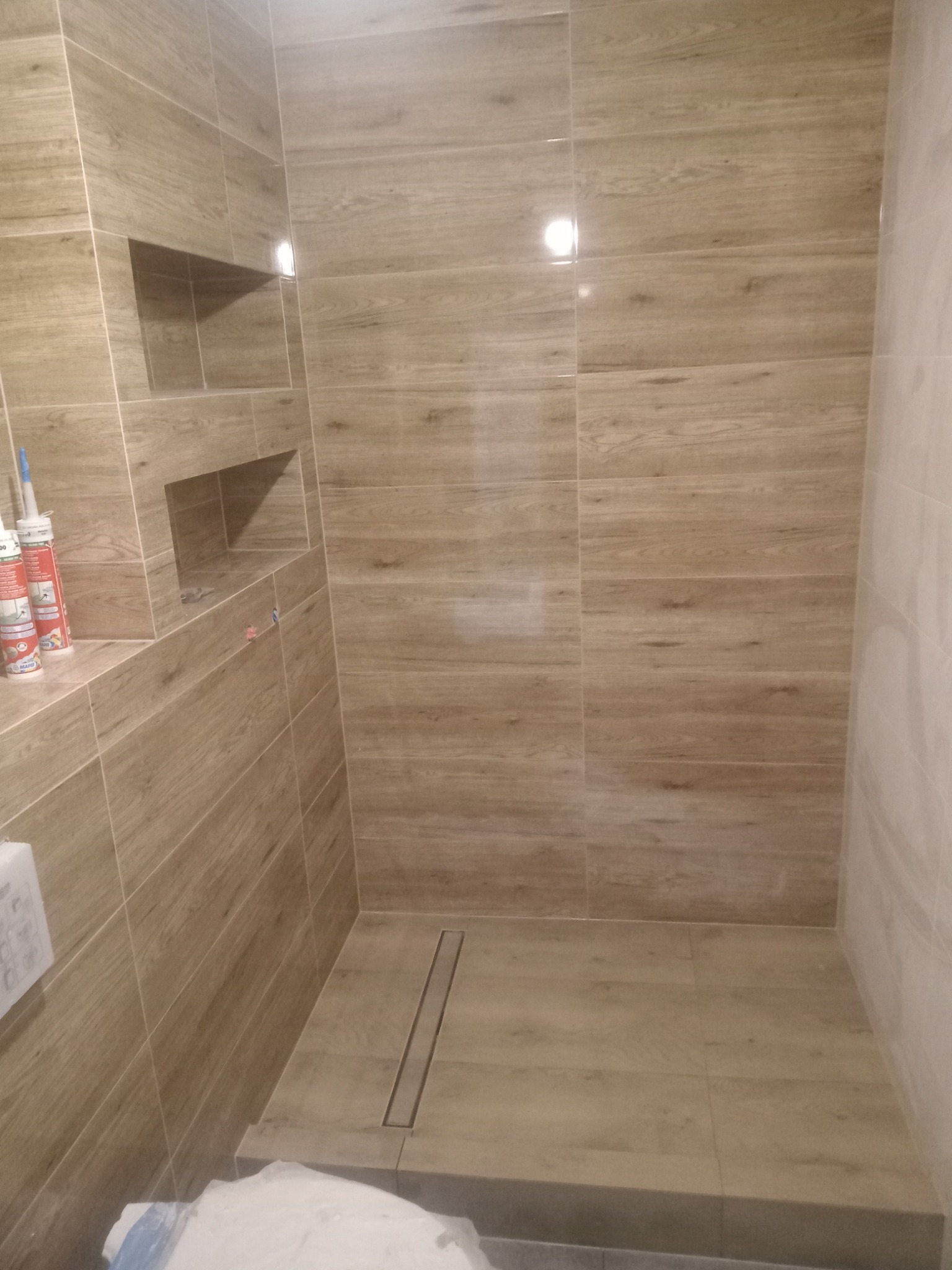 Wykończona kabina prysznicowa z beżowymi płytkami imitującymi drewno, wbudowane półki w ścianie, odpływ liniowy w podłodze, widoczne silikony i akcesoria montażowe na półce.
