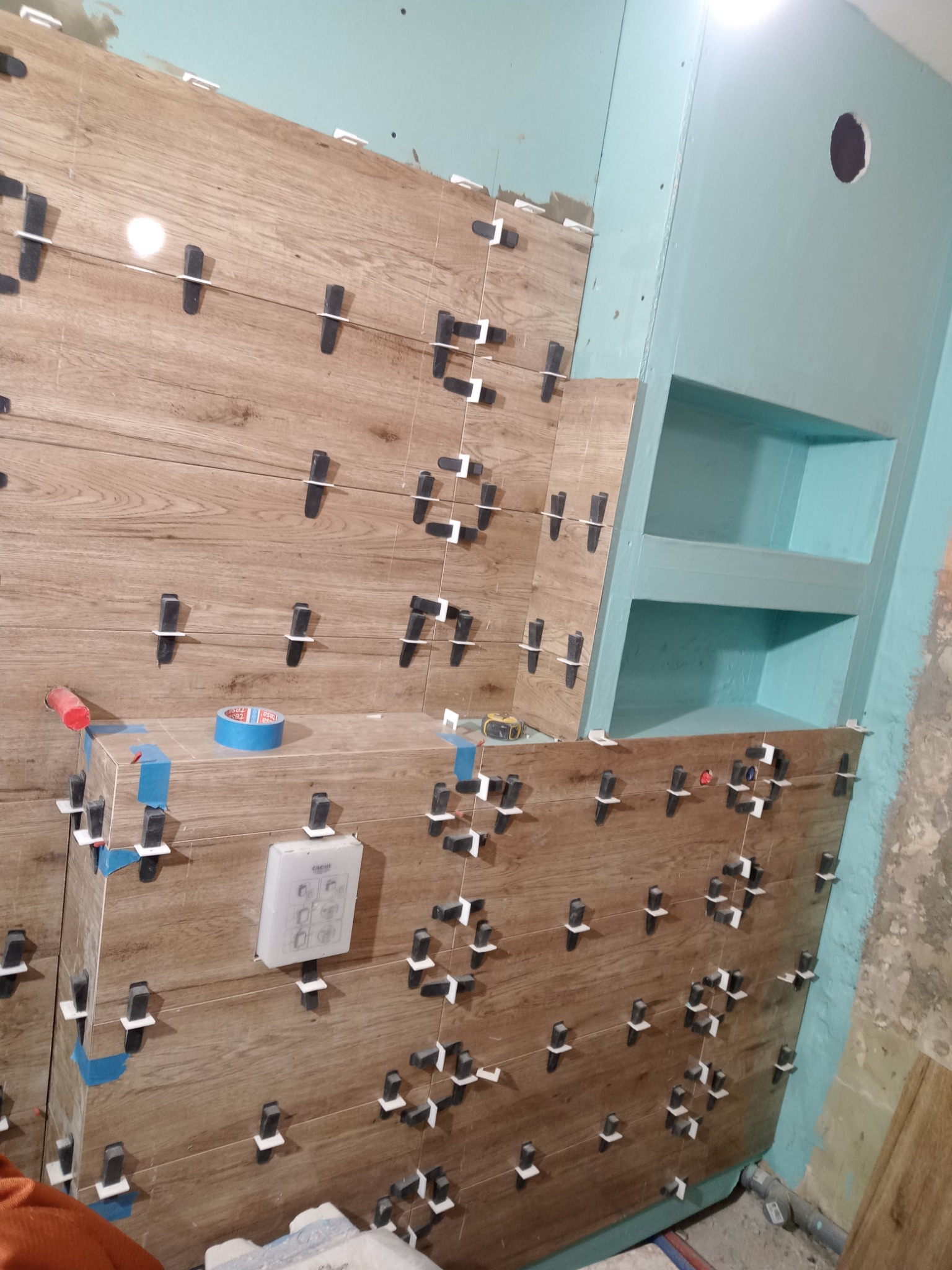 Montaż płytek imitujących drewno na ścianie w łazience, widoczne klipsy poziomujące, fragment nieotynkowanej ściany i wnęka pomalowana na turkusowo.