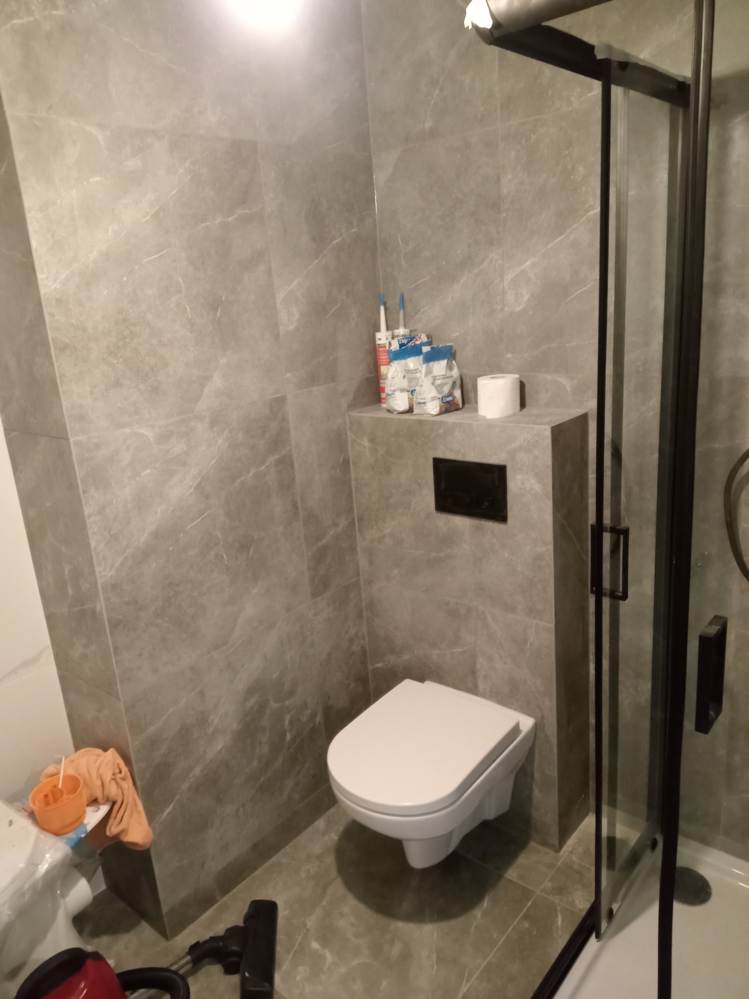 Wykończona łazienka z szarymi płytkami imitującymi marmur, wisząca toaleta z czarnym przyciskiem spłukującym, czarna kabina prysznicowa i materiały budowlane na półce.