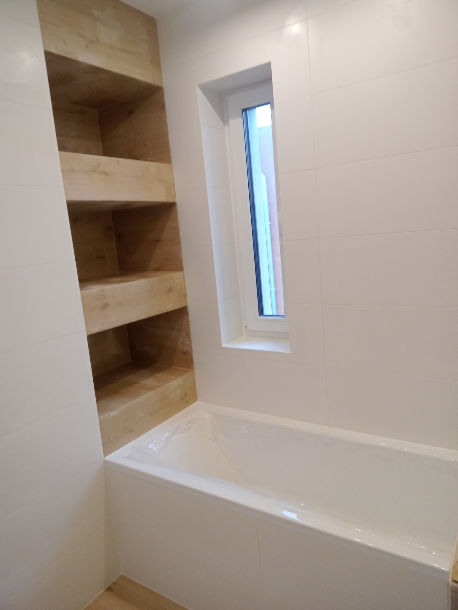 Biała łazienka z wanną, oknem i drewnianą wnęką z półkami, wykończona jasnymi płytkami.