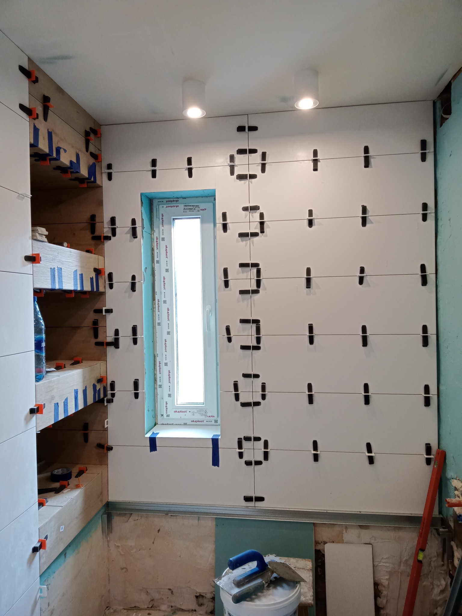 Ściana łazienki w trakcie układania białych płytek, z użyciem poziomych sznurków i klipsów dystansowych, widoczne okno i narzędzia glazurnicze w wiadrze na podłodze.