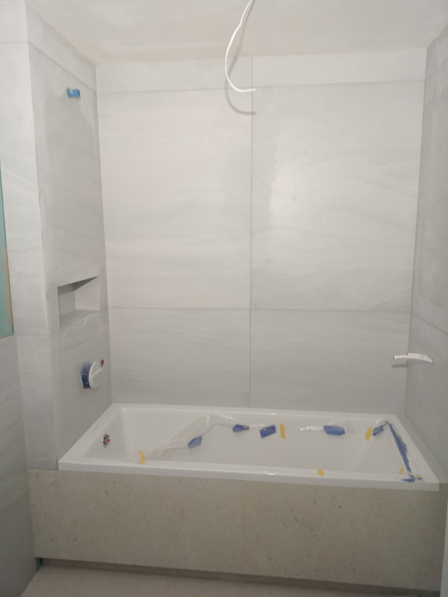 Wanna zabezpieczona folią i taśmą malarską podczas prac wykończeniowych w łazience, widoczne jasne płytki na ścianach z wnęką.
