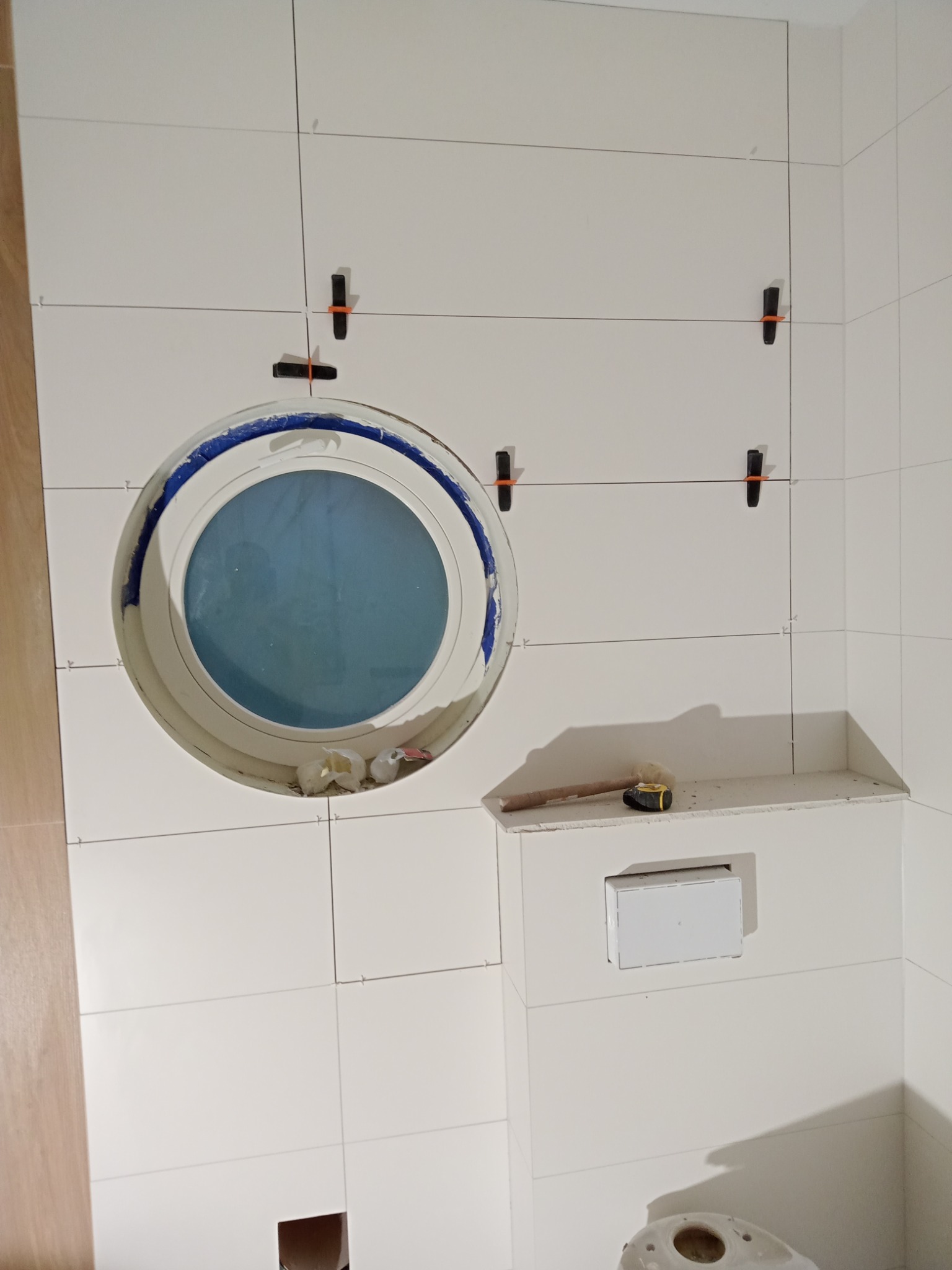 Białe płytki ceramiczne ułożone w łazience, z okrągłym oknem w stylu bulaja, widoczne krzyżyki dystansowe, przymiar i częściowo zainstalowany stelaż podtynkowy WC.