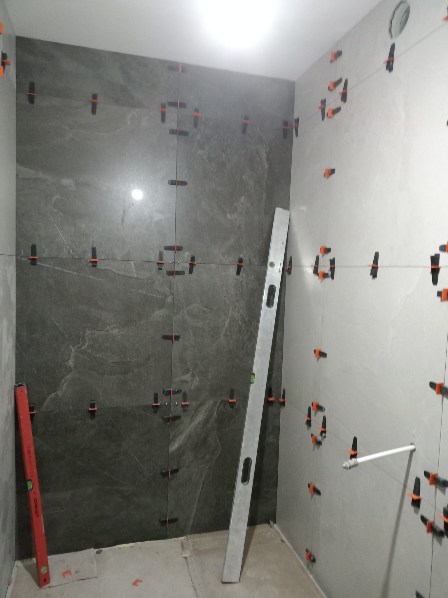 Ściana łazienki w trakcie układania płytek, widoczne poziomice, system poziomowania płytek z czarnymi klipsami i pomarańczowymi klinami, częściowo ułożone płytki w odcieniach szarości.