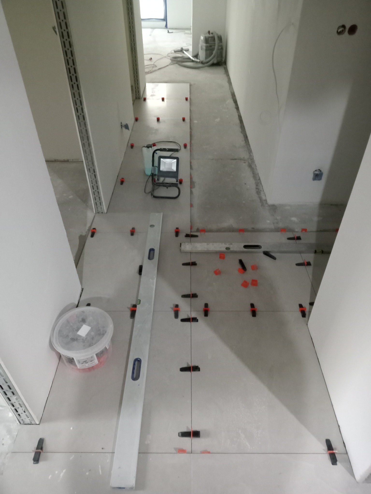 Układanie szarych płytek podłogowych w korytarzu, widoczne poziomica, wiadro z krzyżykami dystansowymi i lampa halogenowa.