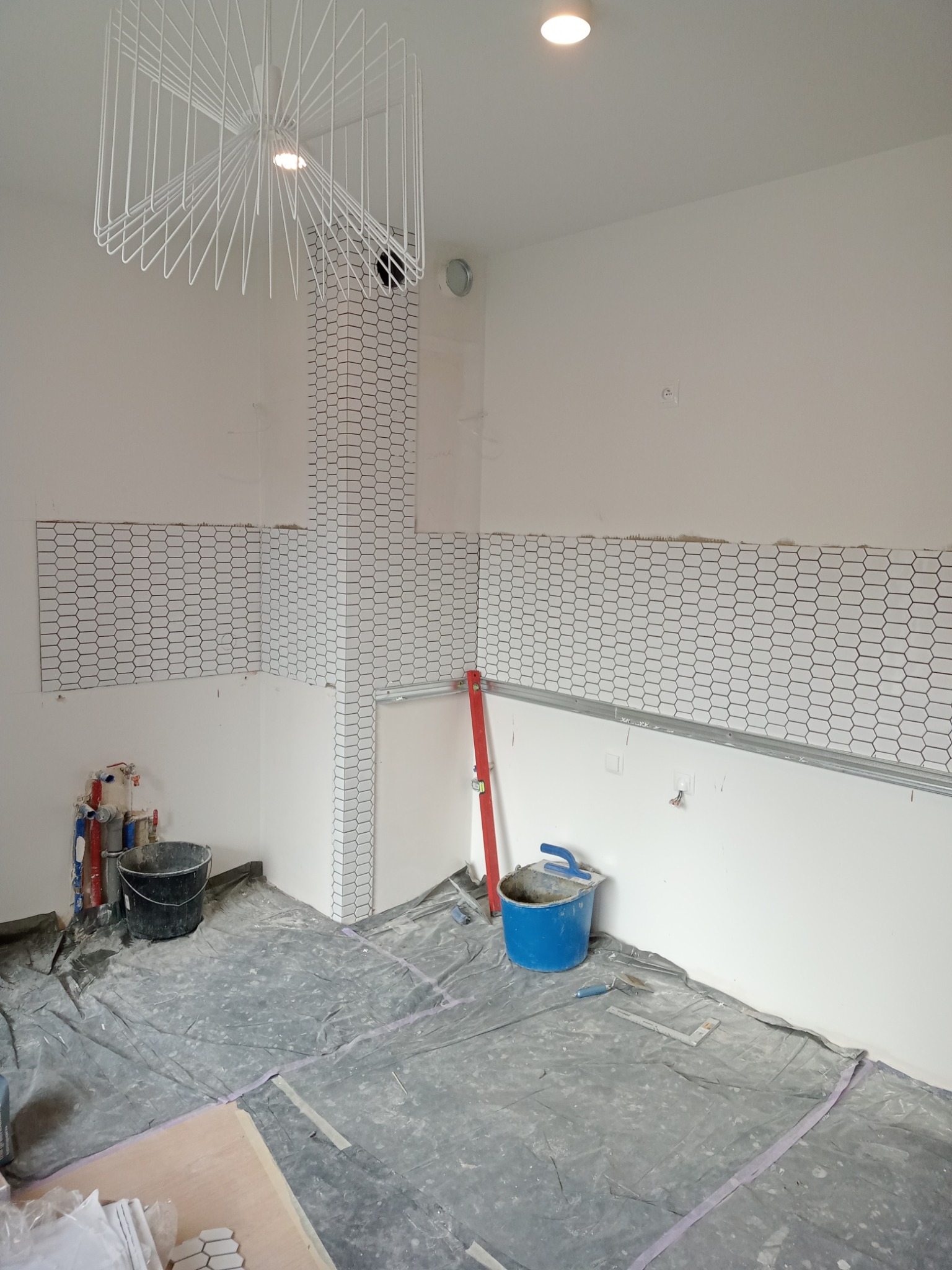 Układanie białych, heksagonalnych płytek na ścianie w trakcie remontu łazienki, widoczne narzędzia glazurnicze, poziomica i wiadro z klejem.