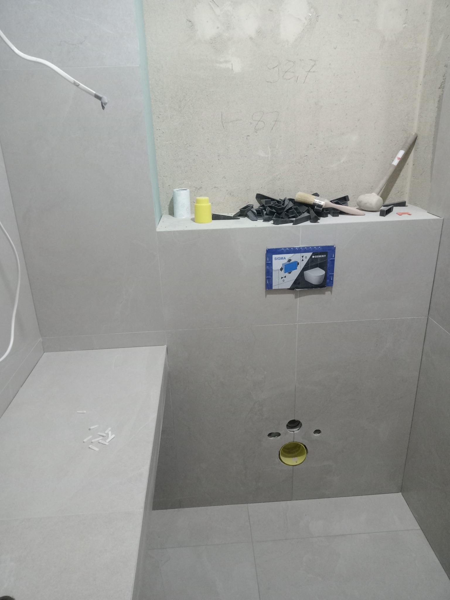 Montaż stelaża podtynkowego Geberit Sigma w łazience wykończonej szarymi płytkami, widoczne narzędzia glazurnicze, poziomica, gumowy młotek i elementy dystansowe na półce nad stelażem.