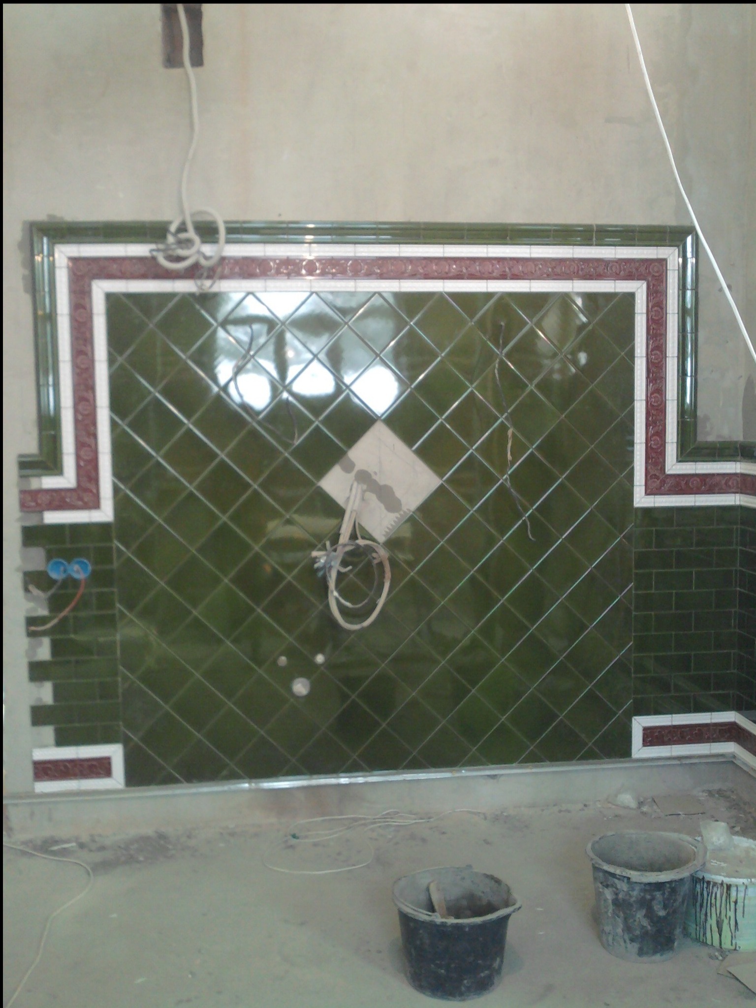 Ściana w trakcie układania zielonej glazury w romby z dekoracyjnym bordiurą, widoczne kable i puszki elektryczne oraz dwa wiadra na podłodze.