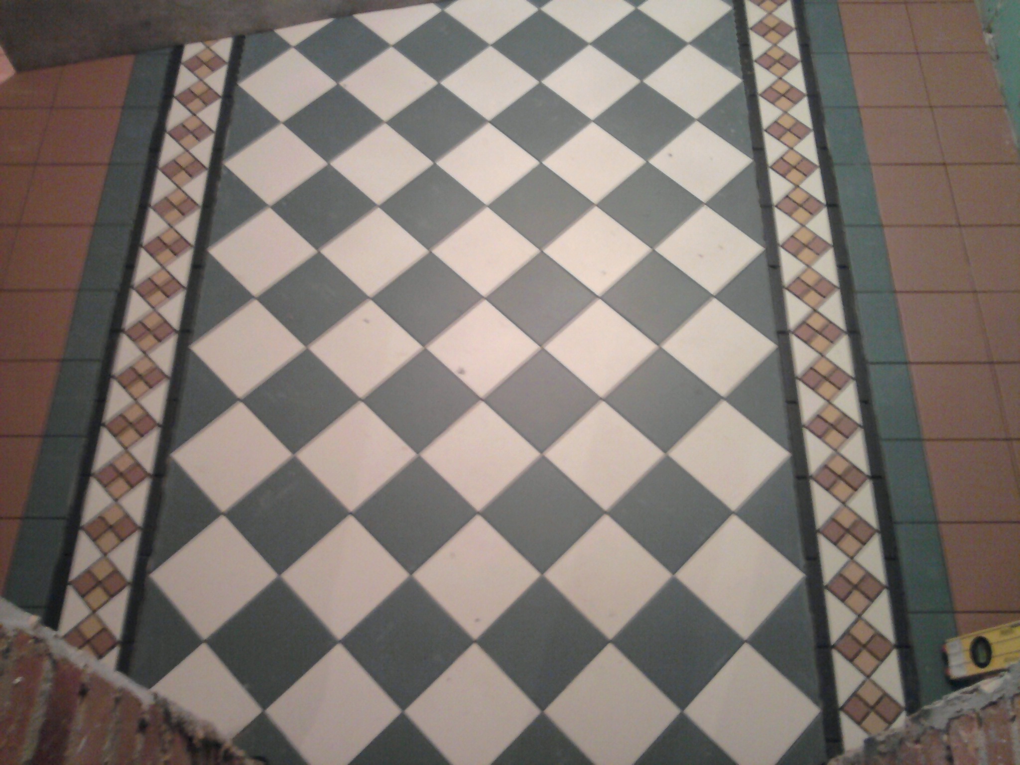 Ułożona podłoga z płytek w szachownicę, z dekoracyjnym pasem brzegowym i poziomnicą w rogu.