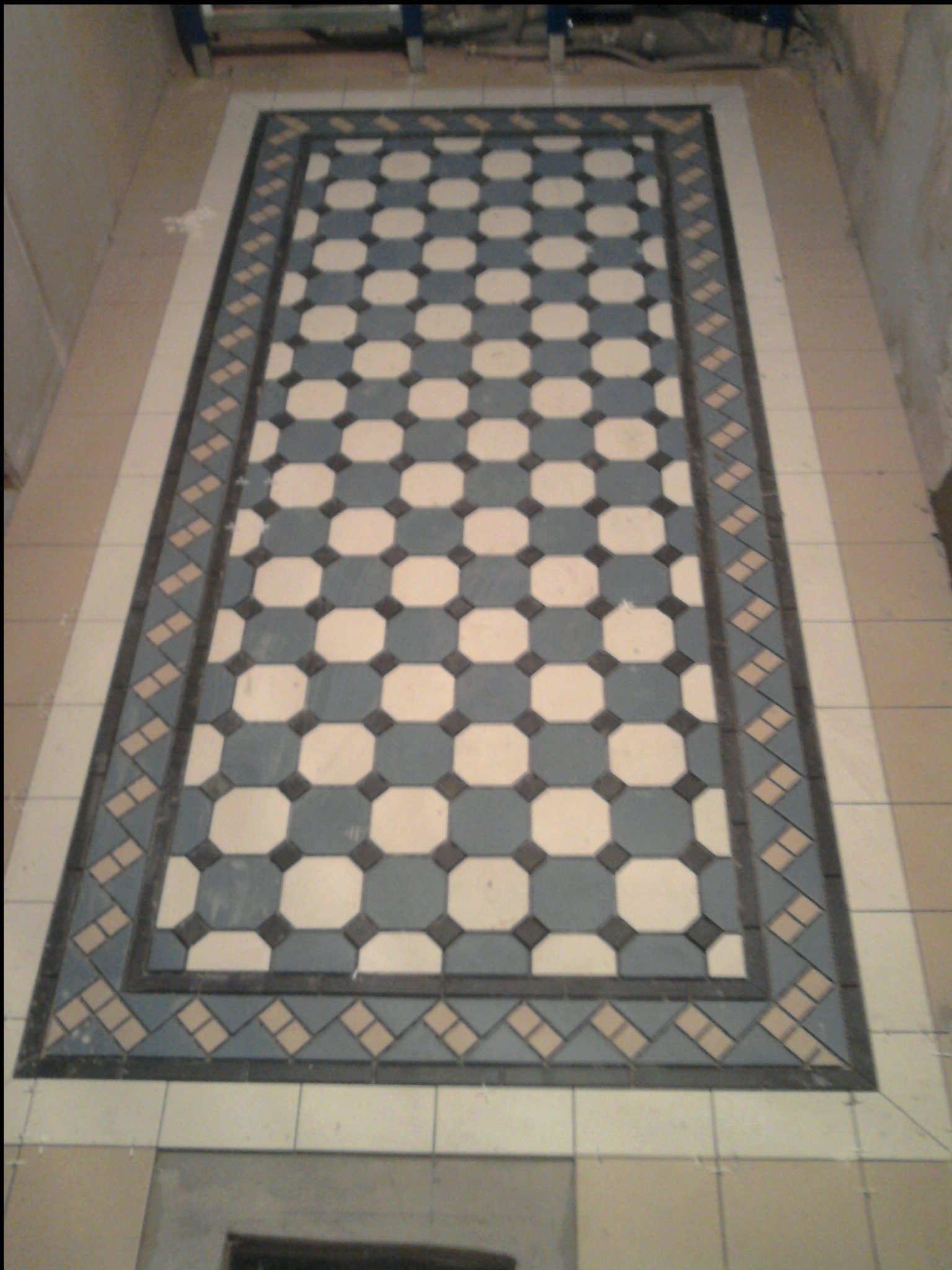 Wzorzysta podłoga z płytek w odcieniach błękitu, bieli i beżu, ułożona w prostokątny dywan z ozdobnym obramowaniem, widok z góry, częściowo zakryta elementami rusztowania.