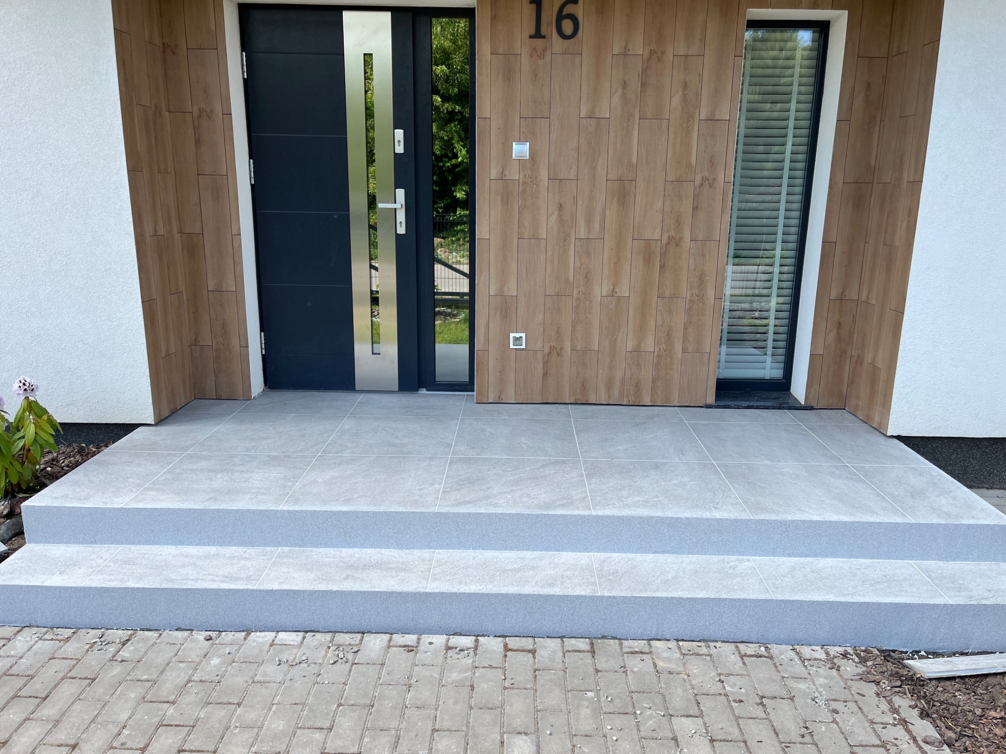 Nowoczesne wejście do domu z szarymi płytkami na schodach i podłodze, czarne drzwi z metalowymi akcentami, numer domu '16' na drewnopodobnej okładzinie elewacyjnej.