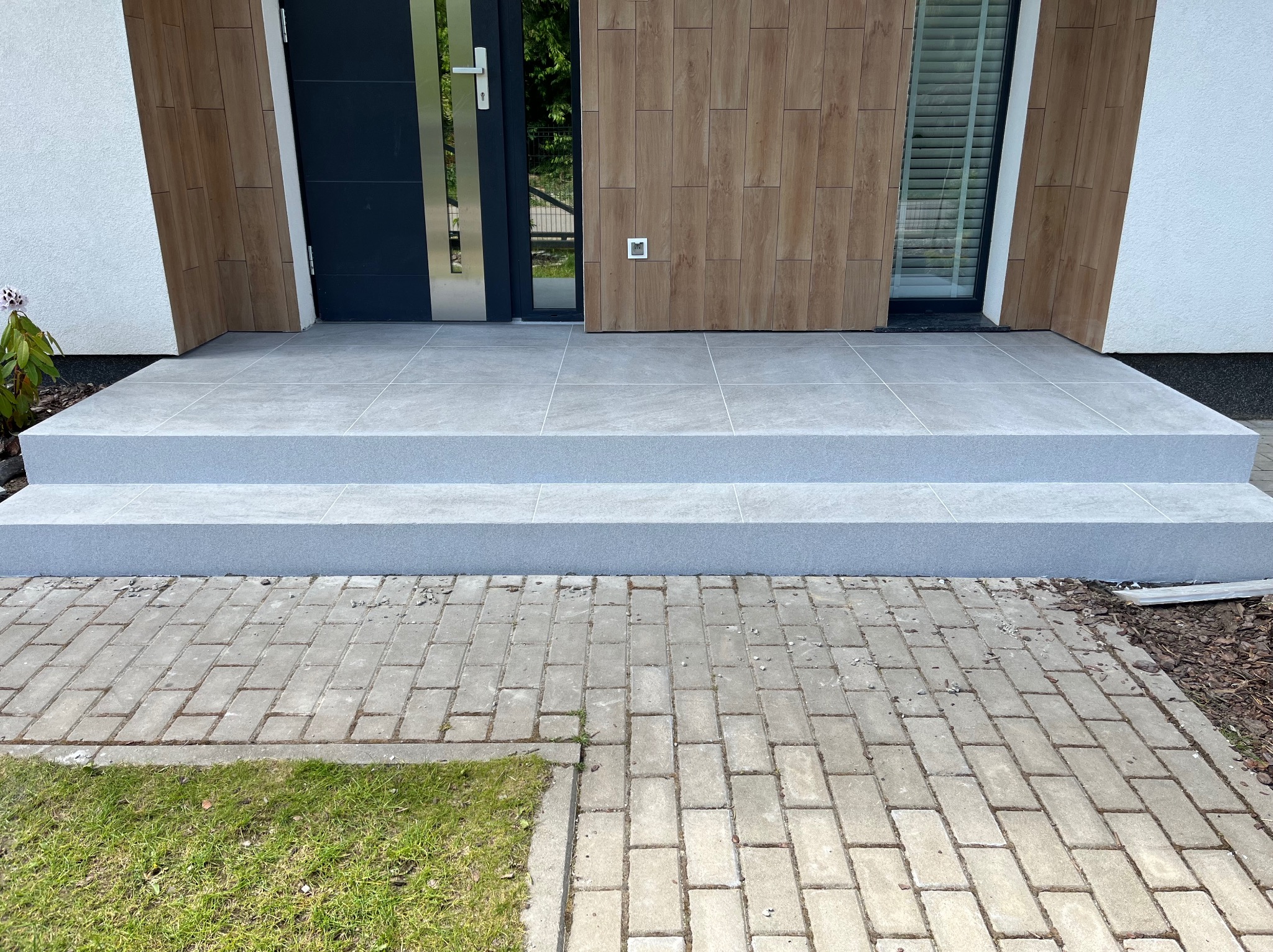 Wyłożone szarymi płytkami schody wejściowe do nowoczesnego domu z drewnianą elewacją i ciemnymi drzwiami, widok z poziomu chodnika z kostki brukowej.