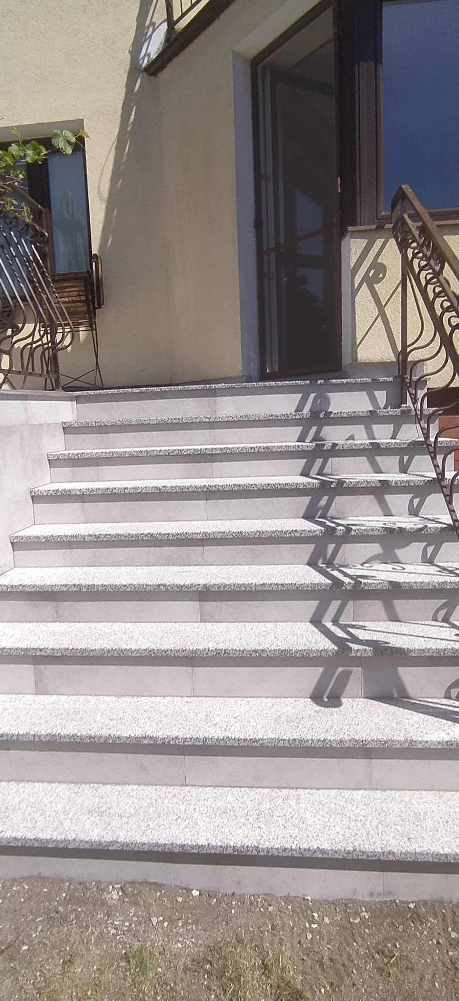 Zewnętrzne schody wyłożone jasnoszarymi płytkami z delikatnym wzorem, prowadzące do drzwi wejściowych z brązową ramą i metalową siatką, z ozdobną metalową balustradą po prawej stronie i widocznym...