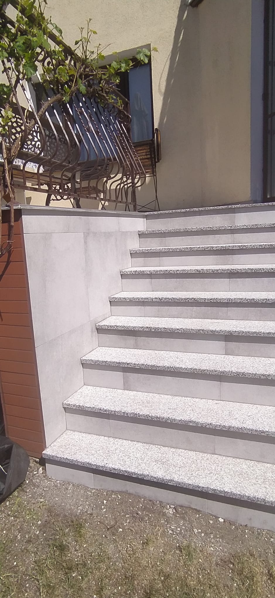 Zewnętrzne schody wyłożone jasnoszarą płytką imitującą kamień, z widocznym fragmentem elewacji budynku i metalowej balustrady balkonu z roślinnością.