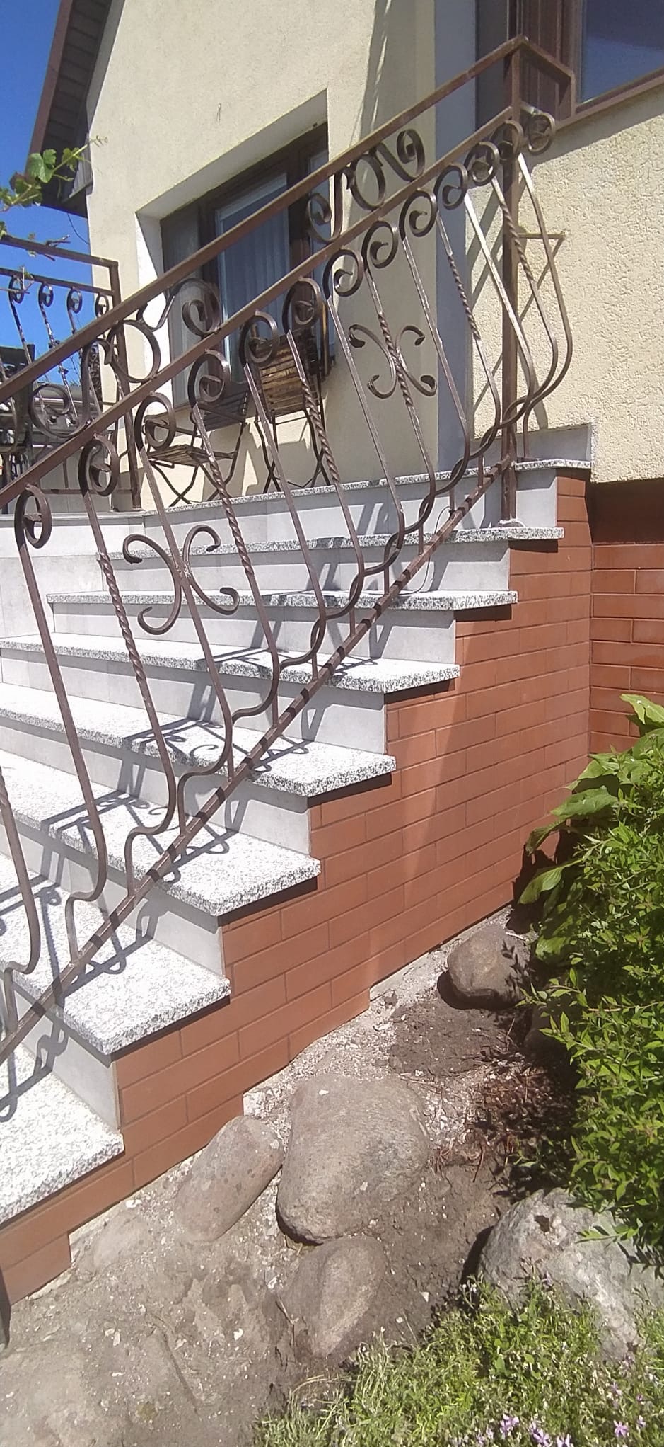 Zewnętrzne schody z szarych płytek granitowych prowadzące do budynku, z ozdobną metalową balustradą i czerwoną cegłą klinkierową na ścianie bocznej.