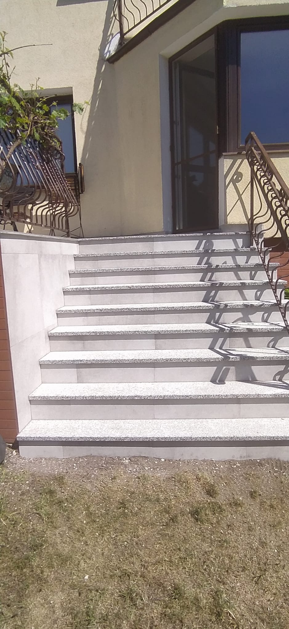Schody zewnętrzne wyłożone jasnoszarymi płytkami z delikatnym wzorem, prowadzące do drzwi wejściowych domu z ozdobną metalową balustradą.