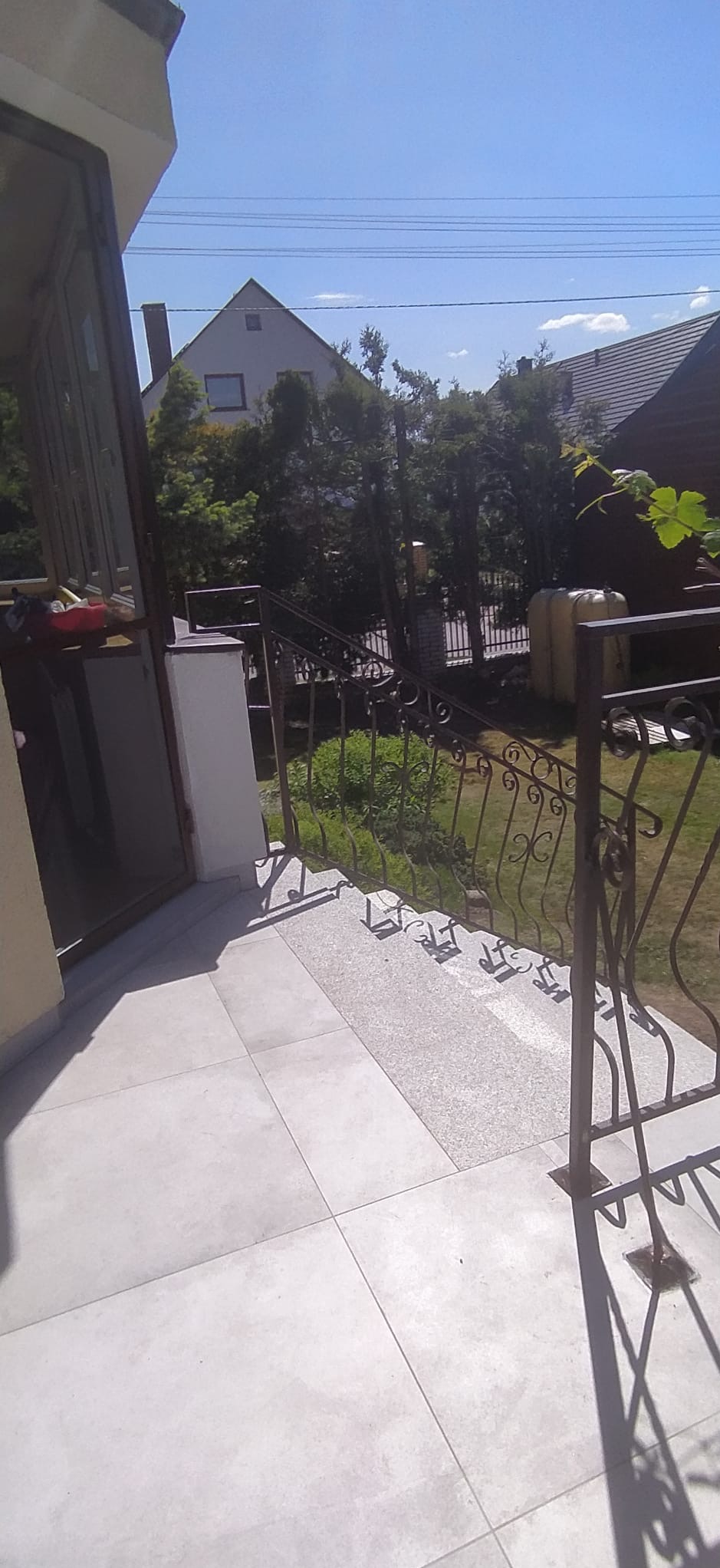 Jasne płytki na tarasie prowadzącym do domu, z metalową, ozdobną balustradą i widokiem na ogród oraz dom w tle.
