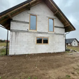 Surowy stan budynku w Kościerzynie z widocznymi oknami z brązowymi ramami, ściany z białych bloczków, przygotowany do dalszych prac wykończeniowych elewacji.