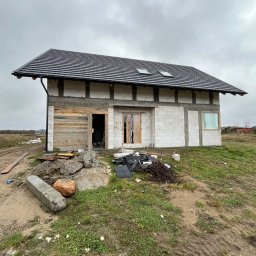 Krystian Zinka - Surowy stan budynku jednorodzinnego z bloczków, z drewnianym szalunkiem na części elewacji i czarnym dachem z lukarnami, na tle pochmurnego nieba.