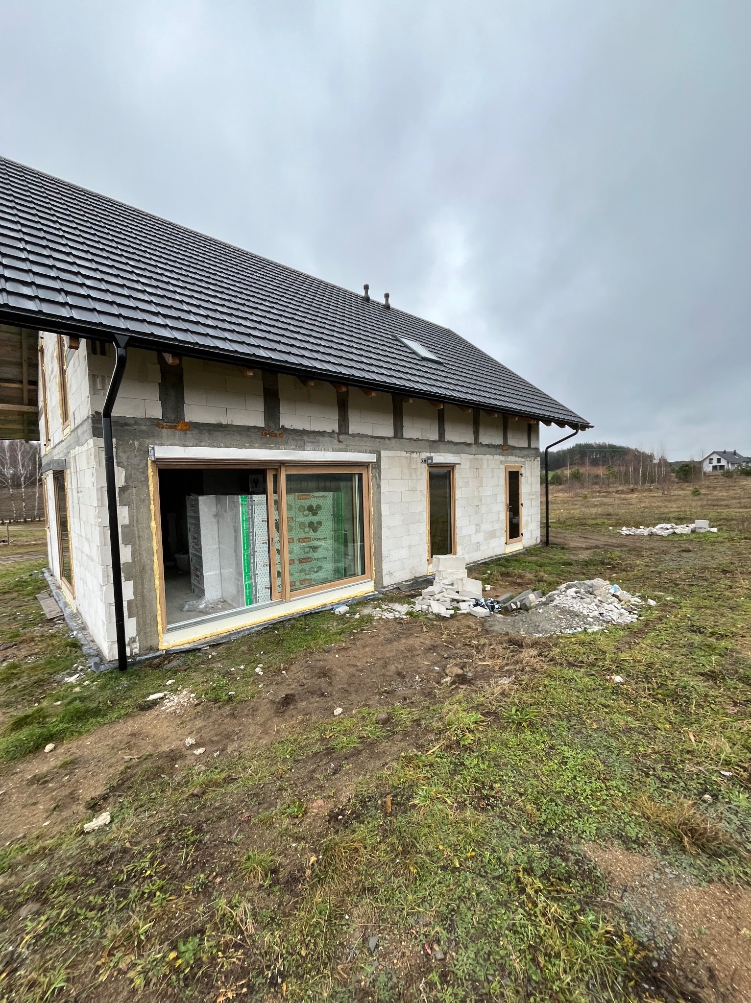 Surowy stan elewacji domu z betonu komórkowego, widoczne okna z drewnianymi ramami, przygotowane do dalszych prac wykończeniowych, teren wokół z resztkami materiałów budowlanych i trawy.