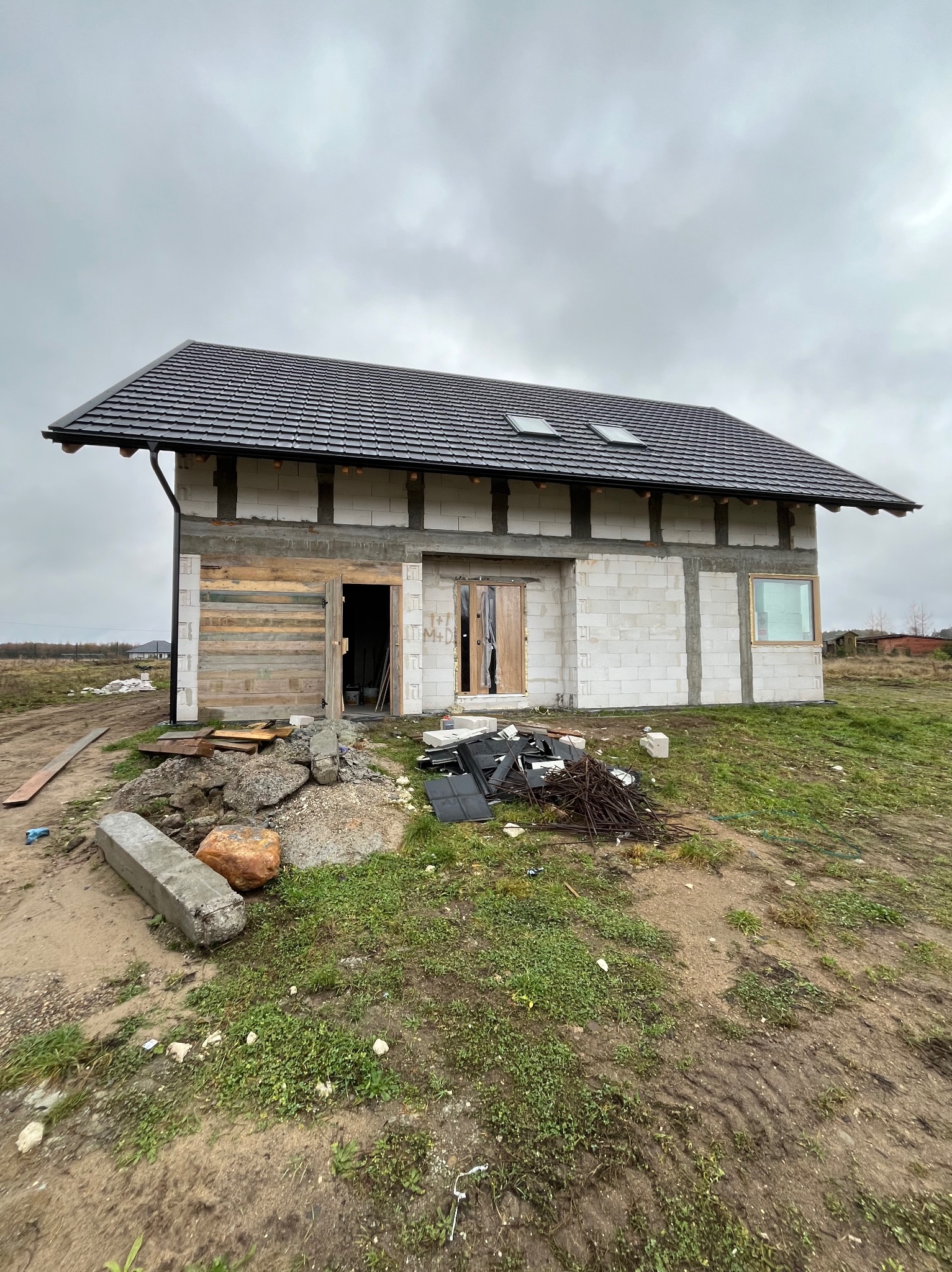 Surowy stan budynku jednorodzinnego z bloczków, z drewnianym szalunkiem na części elewacji i czarnym dachem z lukarnami, na tle pochmurnego nieba.