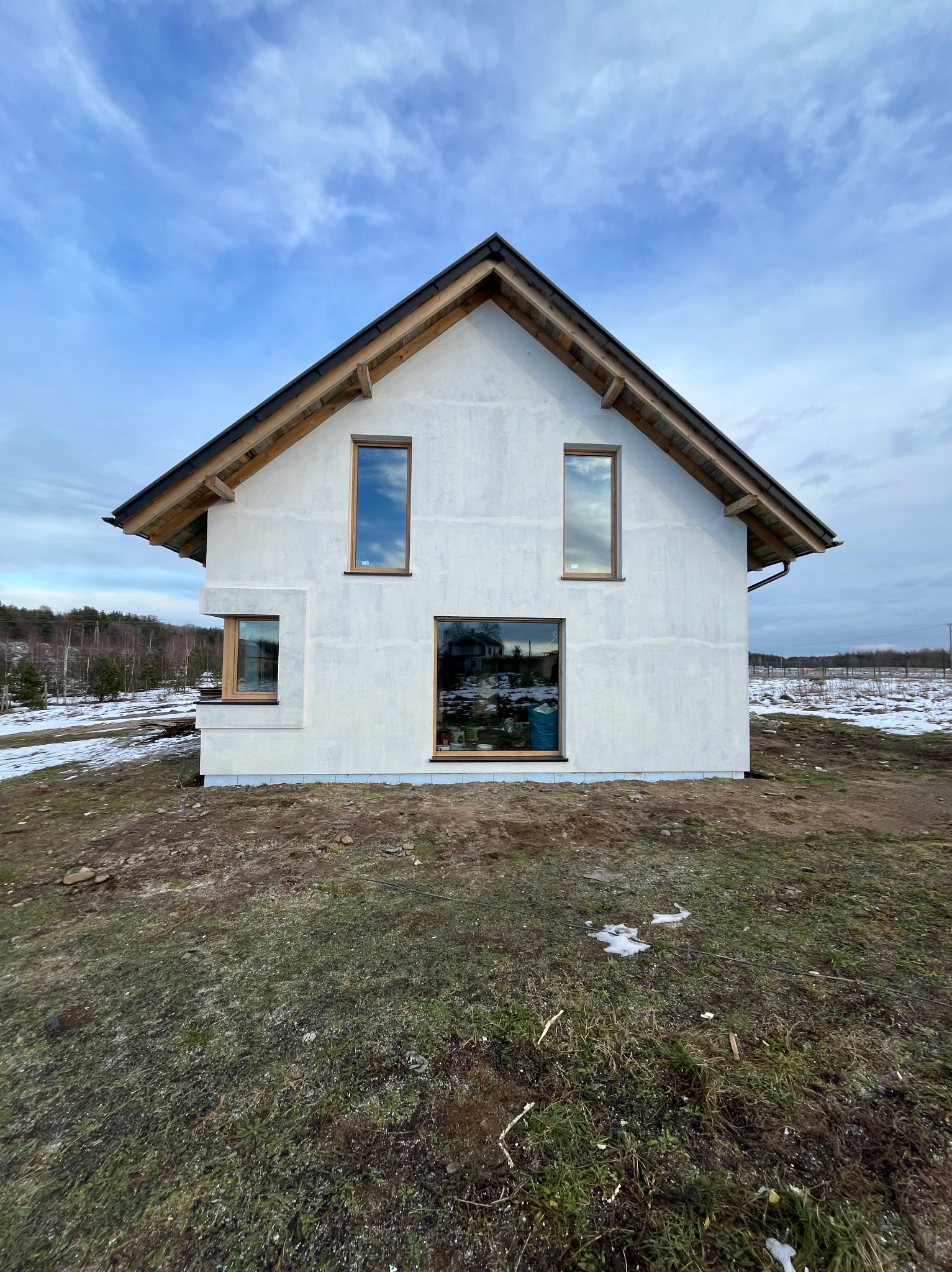 Surowy stan elewacji domu jednorodzinnego z drewnianymi ramami okiennymi, widoczne ślady po nałożeniu tynku, na gruncie resztki śniegu, w tle las i pochmurne niebo.