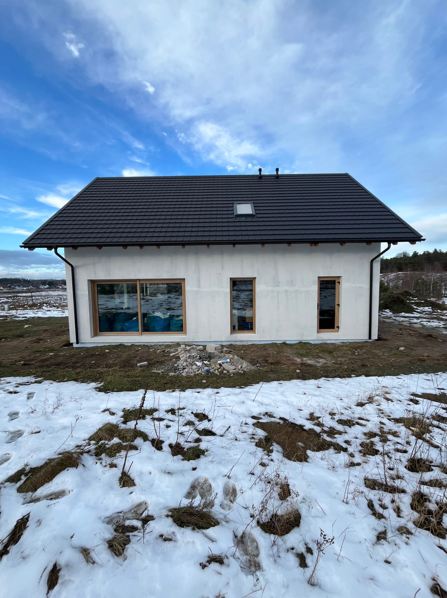 Niedokończony dom jednorodzinny z ocieploną elewacją, widoczne okna bez parapetów, czarna dachówka i fragmenty śniegu na ziemi.